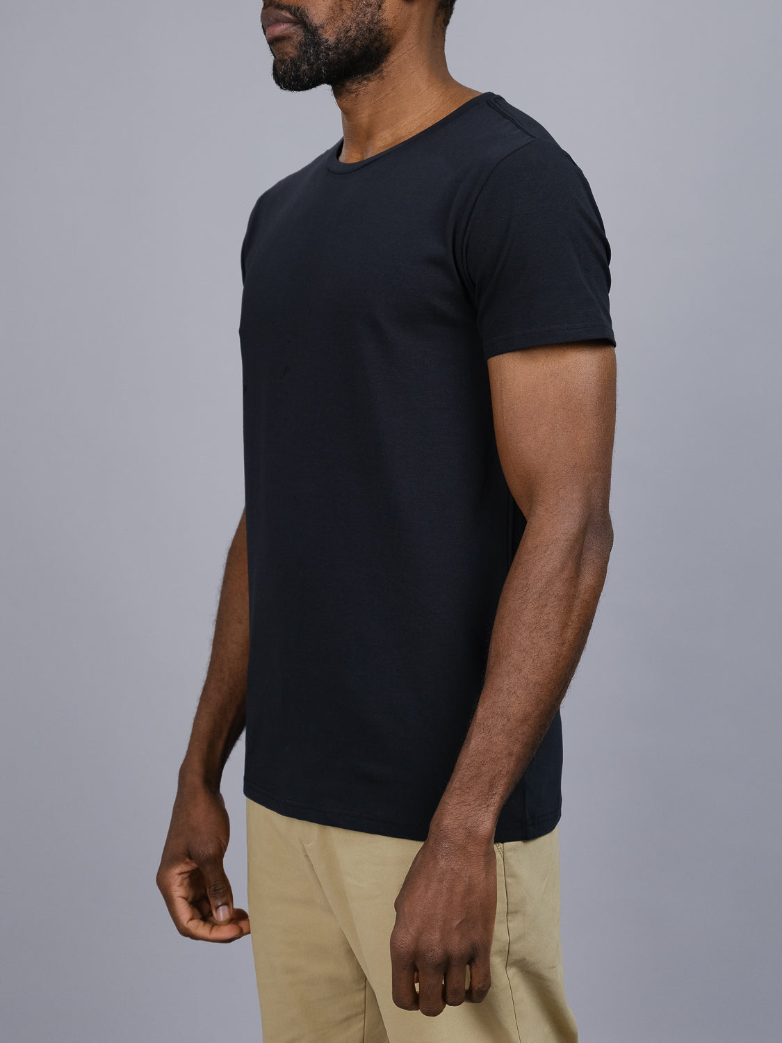 A man in a black ID Menswear Form Crew Neck Tee and khaki pants stands against a gray backdrop. His relaxed posture, with arms at his sides, highlights the form-fitting design and comfortable fabric contrasting the dark tee with the light pants.