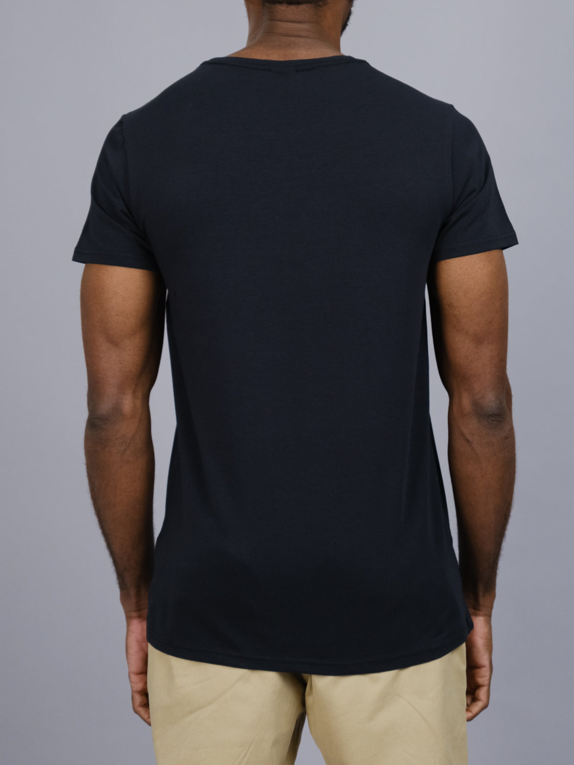 A person is seen from the back wearing ID Menswears Form Crew Neck Tee, a plain black t-shirt made of comfortable fabric, along with beige pants, set against a gray background.