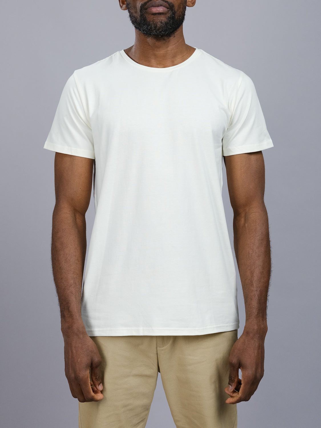 A person wearing ID Menswears Form Crew Neck Tee in plain white with beige pants, made from comfortable fabric, stands against a gray backdrop. The photo is cropped at the neck and knees to highlight the clothing design.