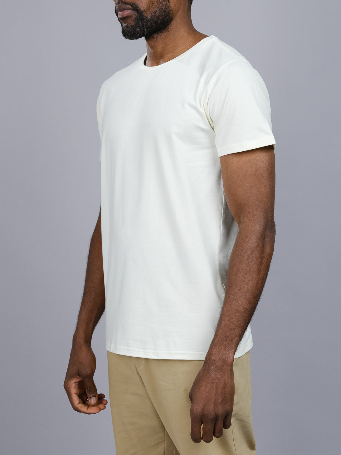 Someone wears the Form Crew Neck Tee and beige pants from ID Menswear against a gray background. The focus is on the torso, with a slightly bent left arm, showcasing the simplicity and form-fitting design of the comfortable fabric.