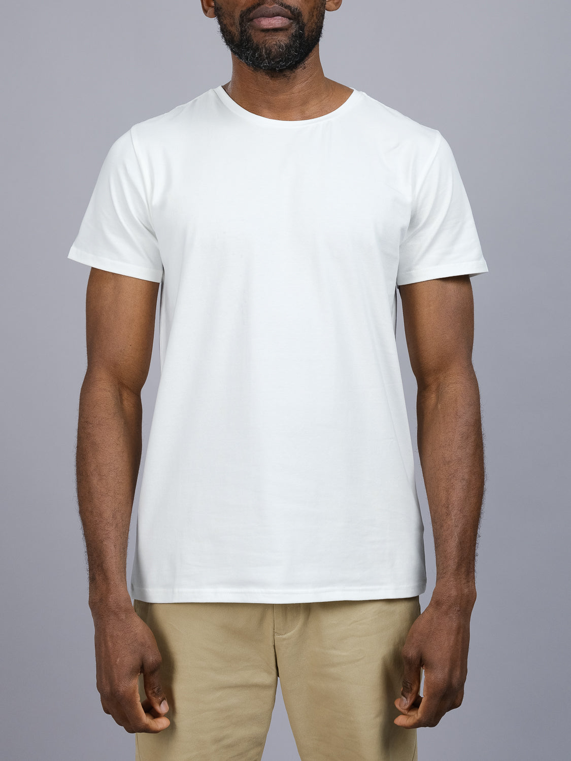 A person in a Form Crew Neck Tee by ID Menswear and beige pants poses against a gray backdrop. The snug fit on the torso and upper legs, with relaxed arms at their sides, embodies minimalist elegance.