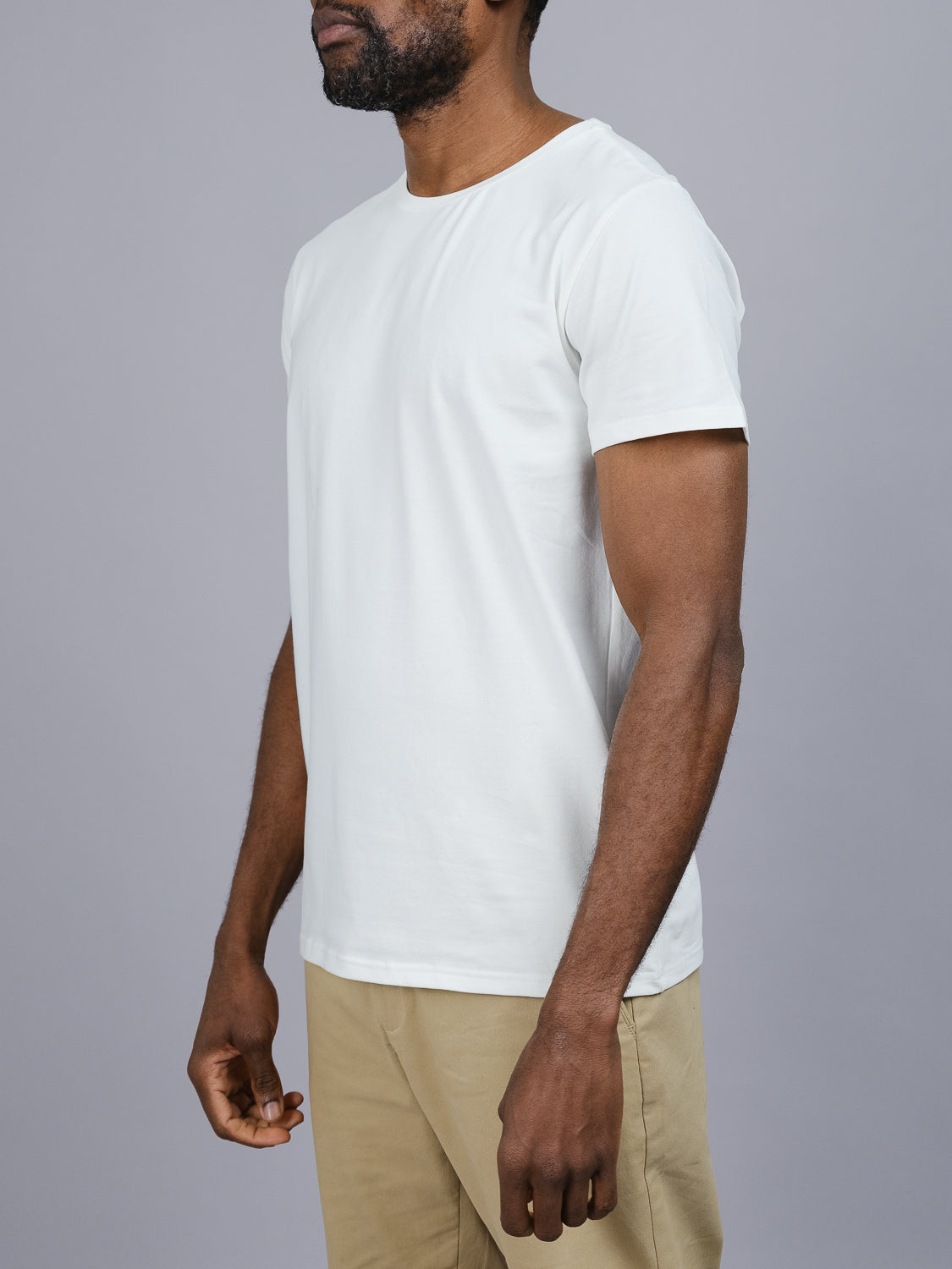 A person wearing an ID Menswear Form Crew Neck Tee in plain white and beige pants stands against a gray background. The form-fitting design enhances their side-facing pose with arms relaxed by their sides.