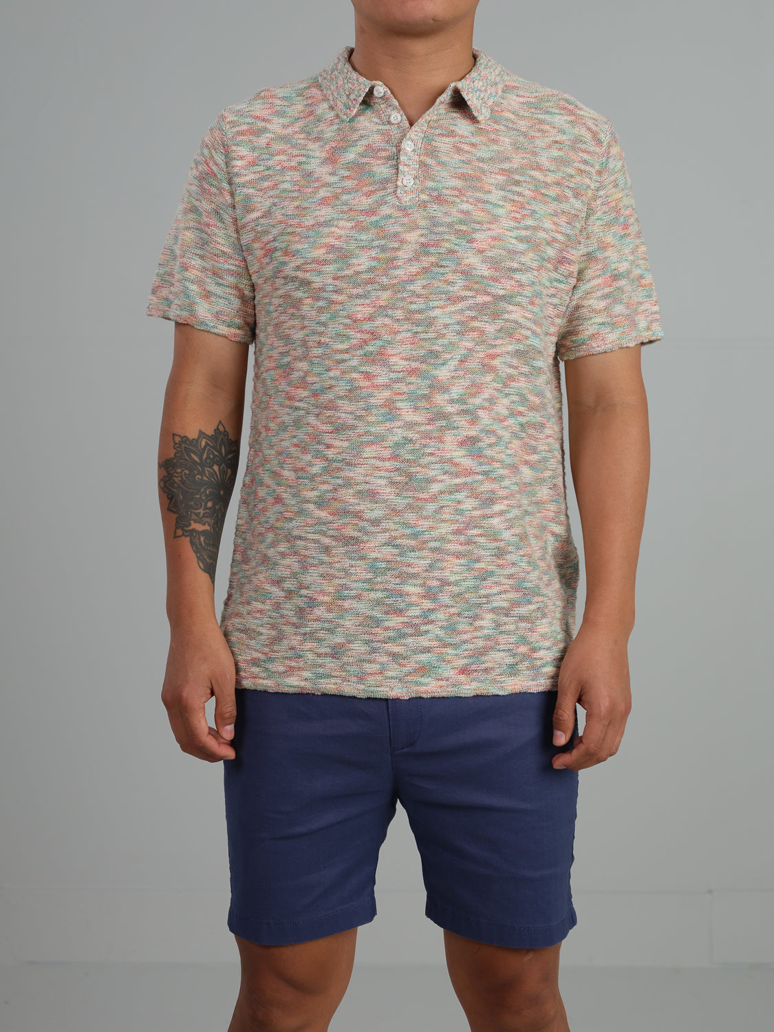 A person in an ID Menswear Miami Reverse Knit Cotton Polo, featuring a multicolored design and short sleeves, pairs it with blue shorts against a plain background. The breathable fit elevates their casual vibe, revealing a tattoo on their left forearm.