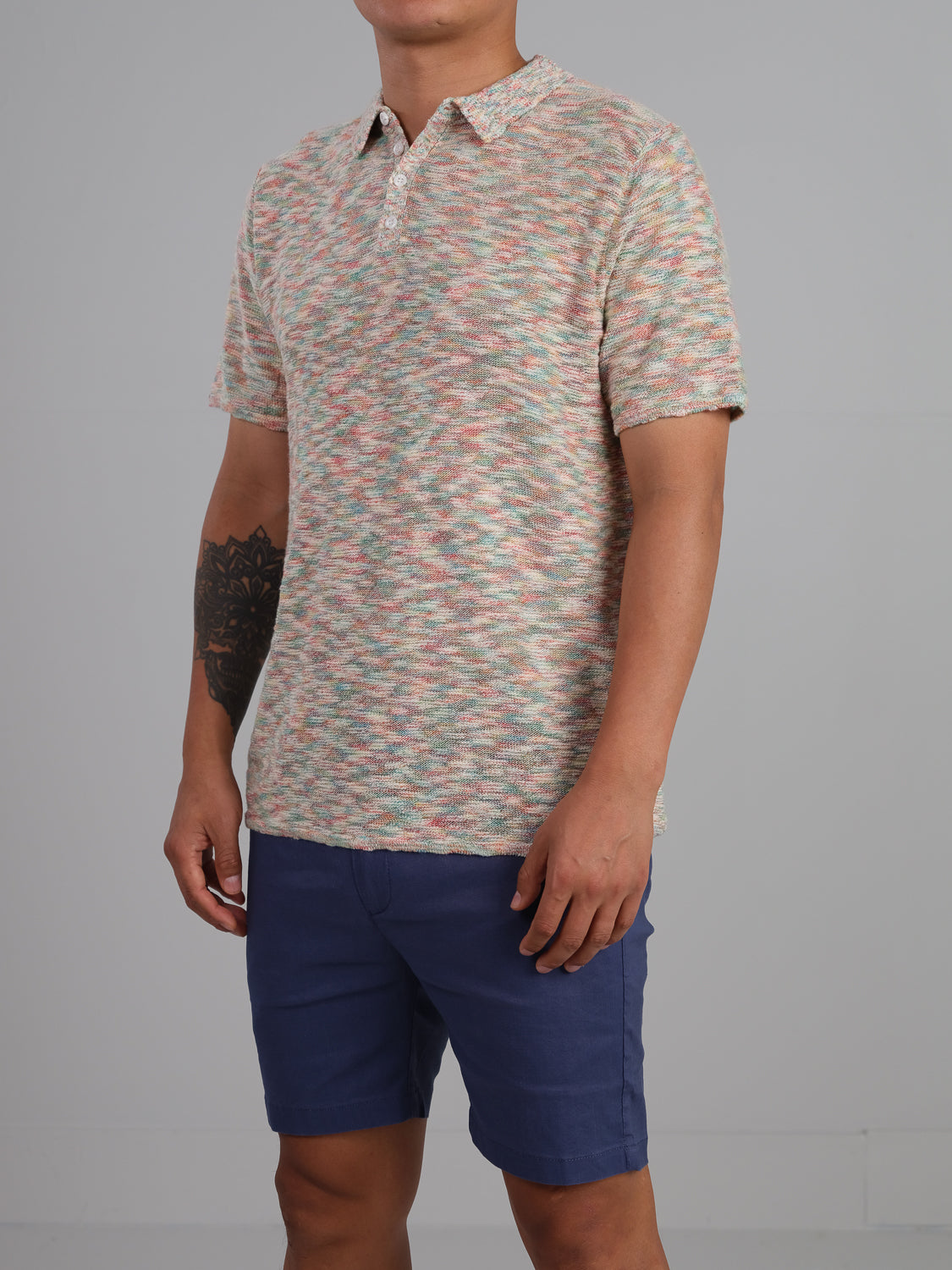 Sporting a Miami Reverse Knit Cotton Polo by ID Menswear, a person embraces relaxed casual wear with this multicolored, patterned short-sleeve polo and blue shorts. Their left forearm tattoo complements the plain neutral backdrop.