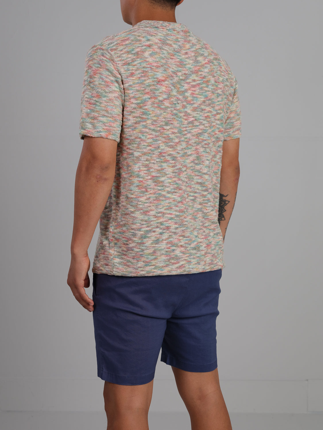 A person wearing the Miami Reverse Knit Cotton Polo by ID Menswear and blue shorts stands against a plain gray background, showcasing a tattoo on their left forearm as a unique touch to their casual ensemble.