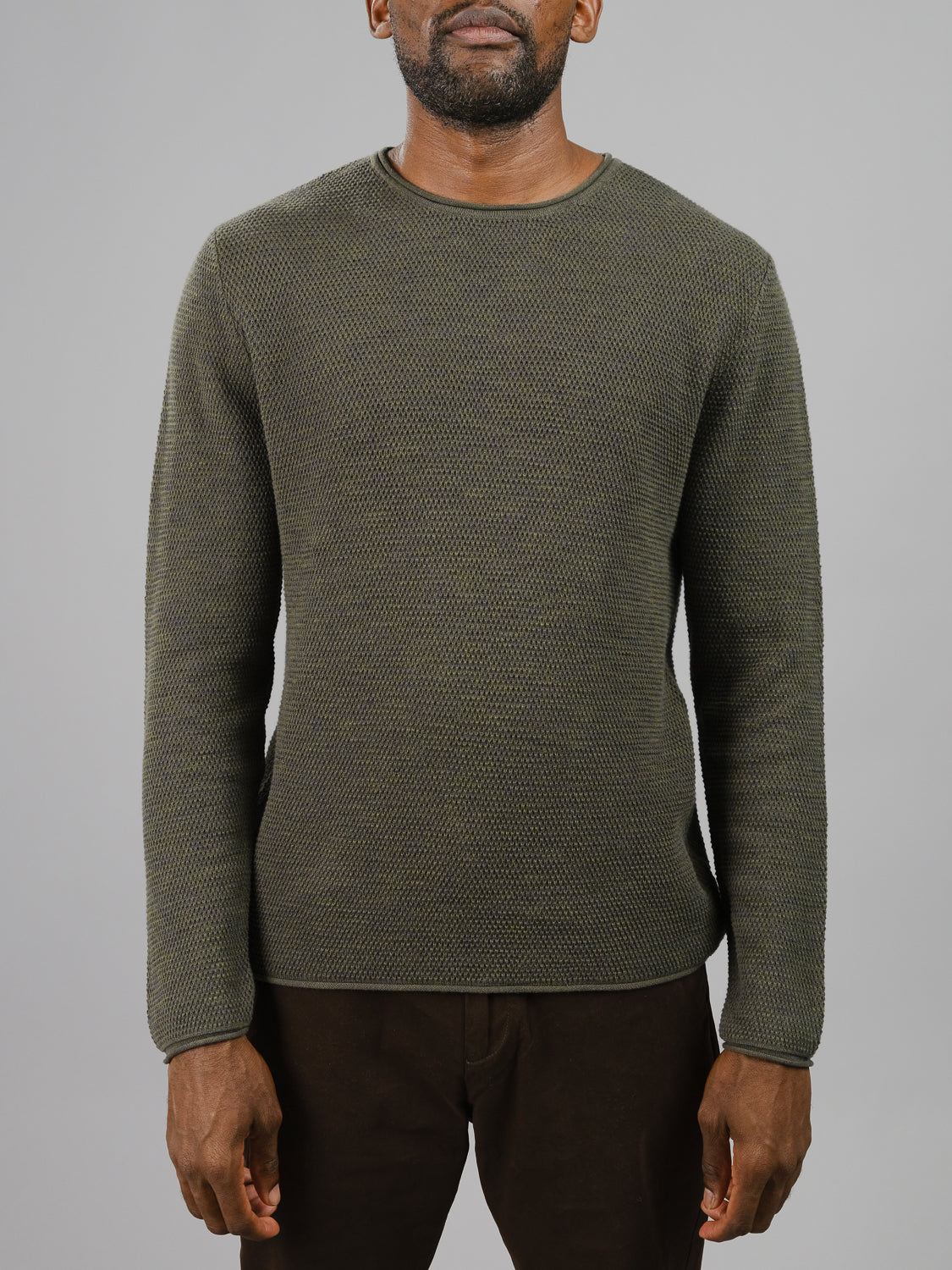 A man in an ID Menswear Malta Rolled Neck Sweater Tee, featuring a textured olive green and comfortable fit, paired with brown pants, stands against a plain gray background. His face is not visible in the image.