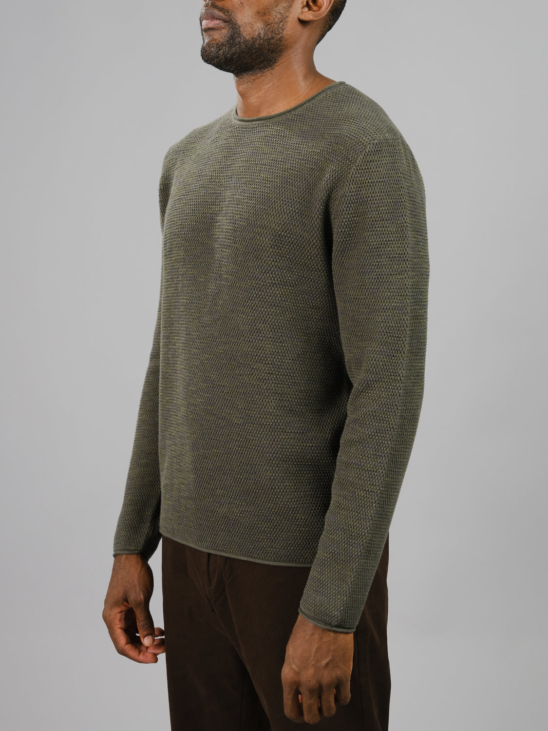 A person is seen against a plain gray background wearing textured green Malta Rolled Neck Sweater Tee by ID Menswear and brown pants, with arms at their sides. The soft cotton tee provides a comfortable fit, but the persons head is not visible.