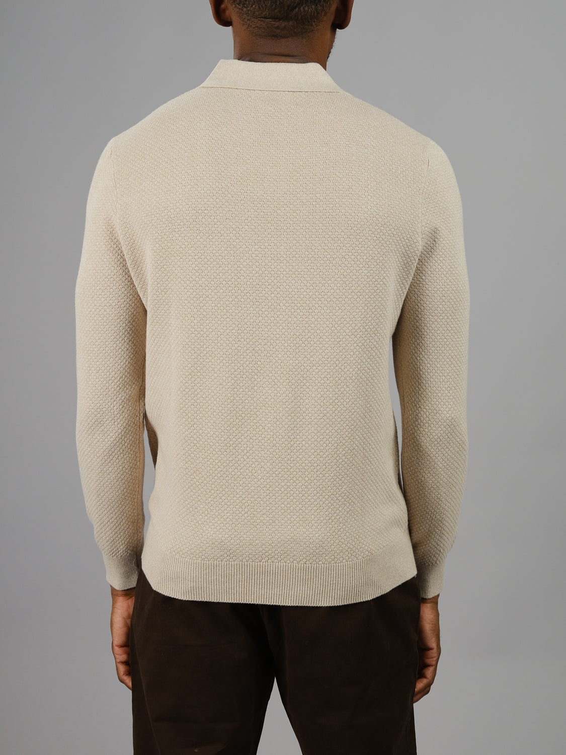A person wearing the Seattle Long Sleeve Cotton Polo by ID Menswear faces away, highlighting its subtle textured pattern. Paired with dark brown pants against a plain gray background, this beige polo is perfect for smart-casual occasions, exuding understated elegance.