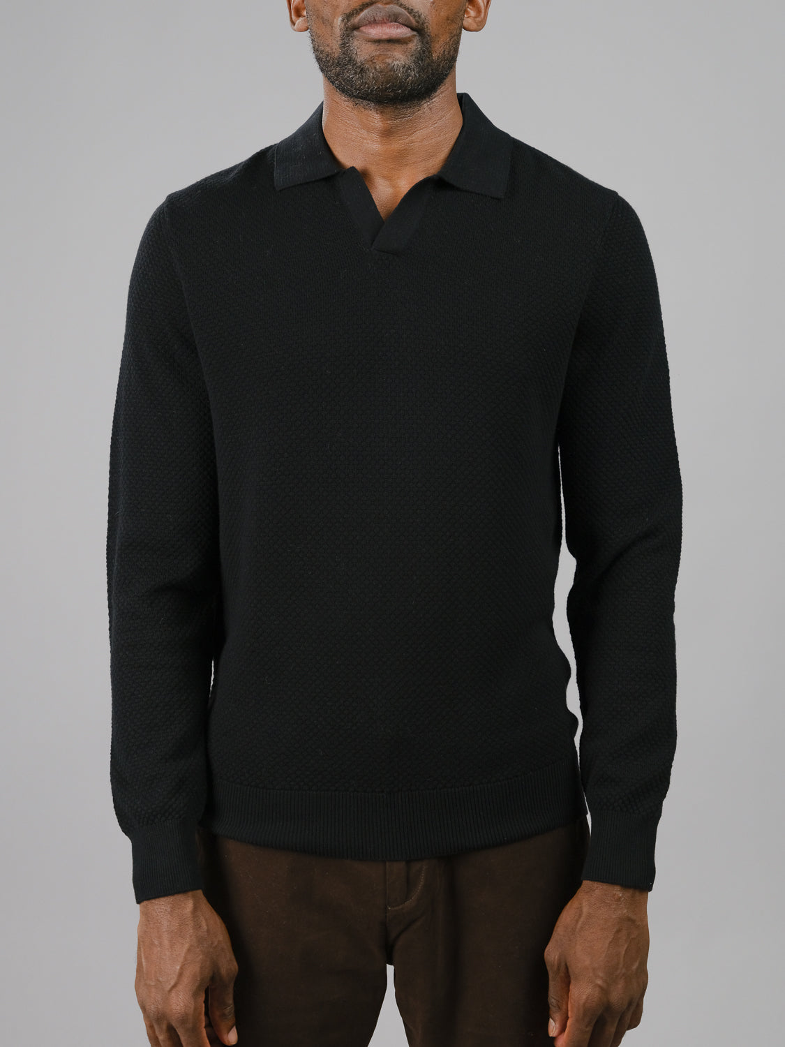 A man in a textured black Seattle Long Sleeve Cotton Polo by ID Menswear stands against a gray backdrop, perfect for smart-casual settings. With short hair and a beard, hes styled with dark pants for an effortlessly polished and relaxed appearance.
