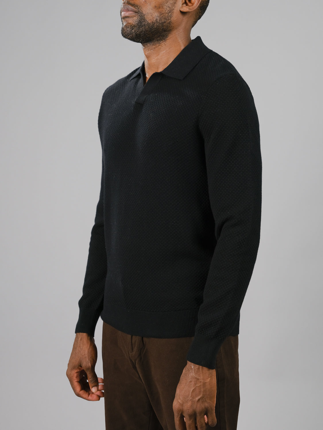 A man stands against a gray backdrop wearing ID Menswears black Seattle Long Sleeve Cotton Polo and brown pants. The focus is on his smart-casual attire as his head is cropped out of the frame.