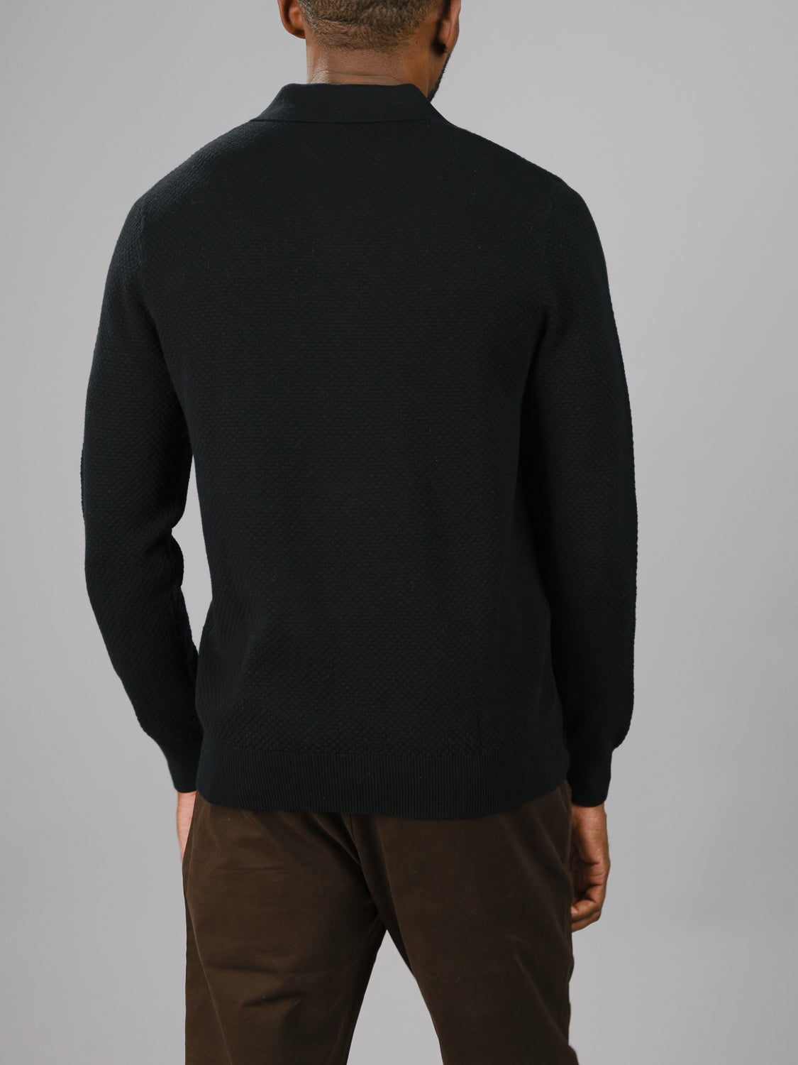 A person wearing the black Seattle Long Sleeve Cotton Polo by ID Menswear stands against a plain gray background, paired with brown pants, exuding an effortless vibe ideal for smart-casual occasions.