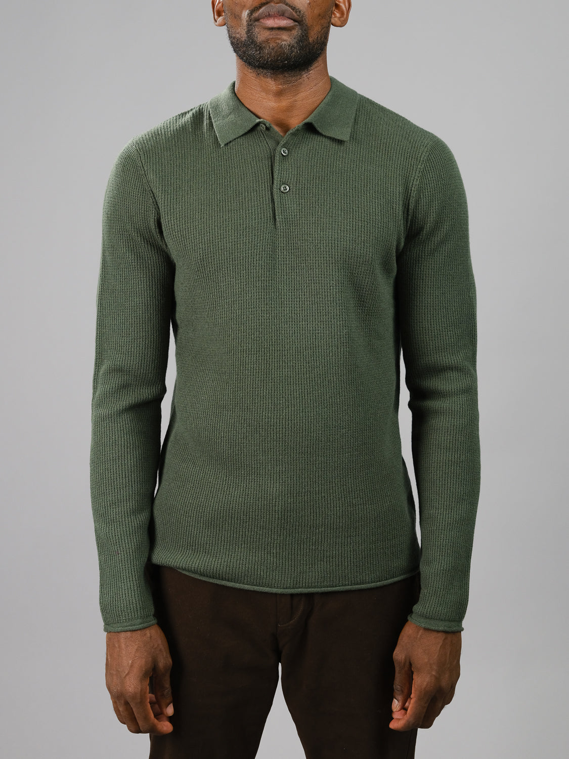 Wearing the Gothenburg Merino Sweater Polo by ID Menswear in dark green, a person embodies casual elegance with its long sleeves and button-up collar, paired with dark pants against a plain gray backdrop.