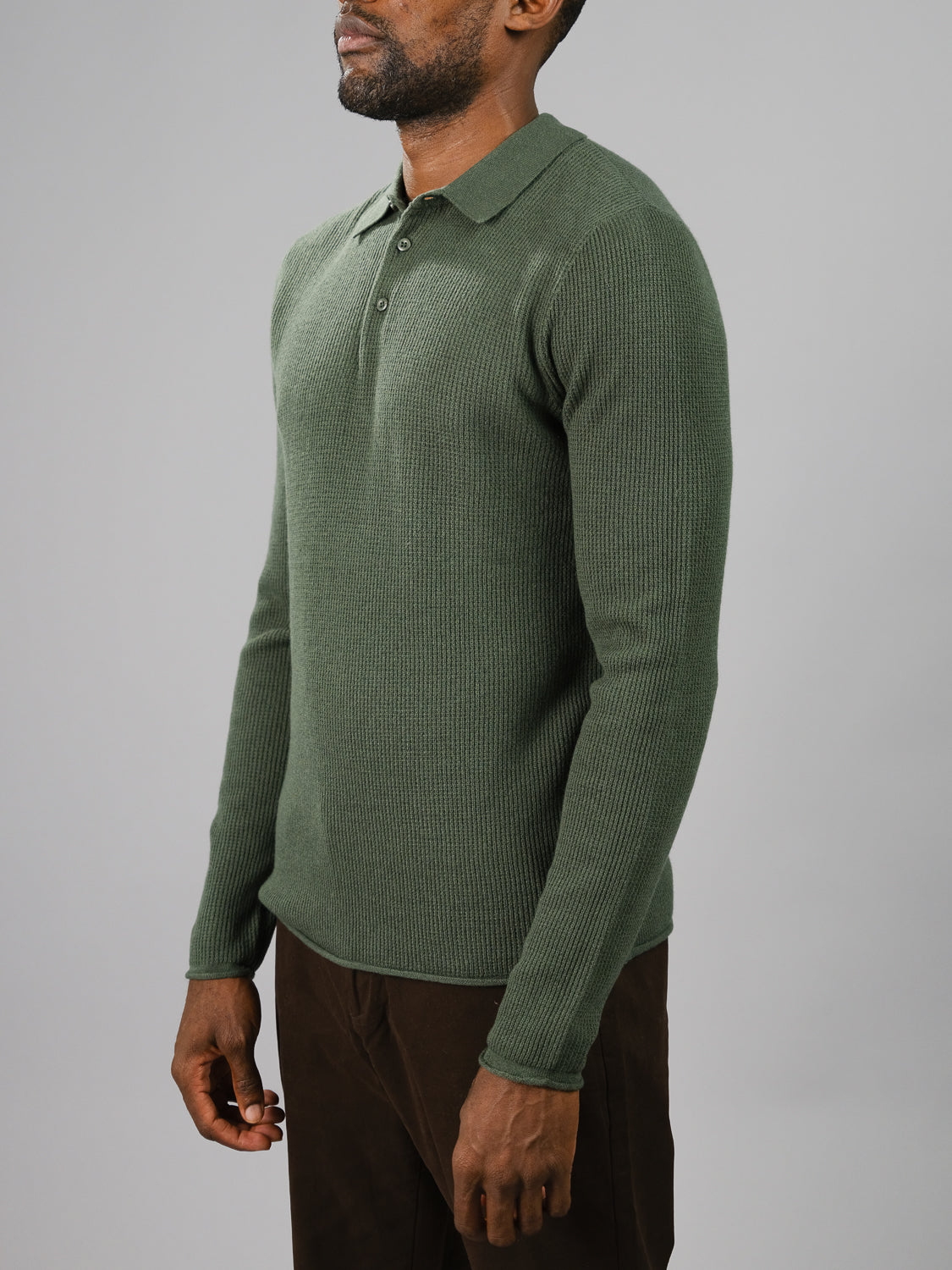Wearing the Gothenberg Merino Sweater Polo by ID Menswear, a person exudes casual elegance in green ribbed fabric against a plain gray background. Dark pants and a relaxed posture beautifully highlight the shirts texture and style.