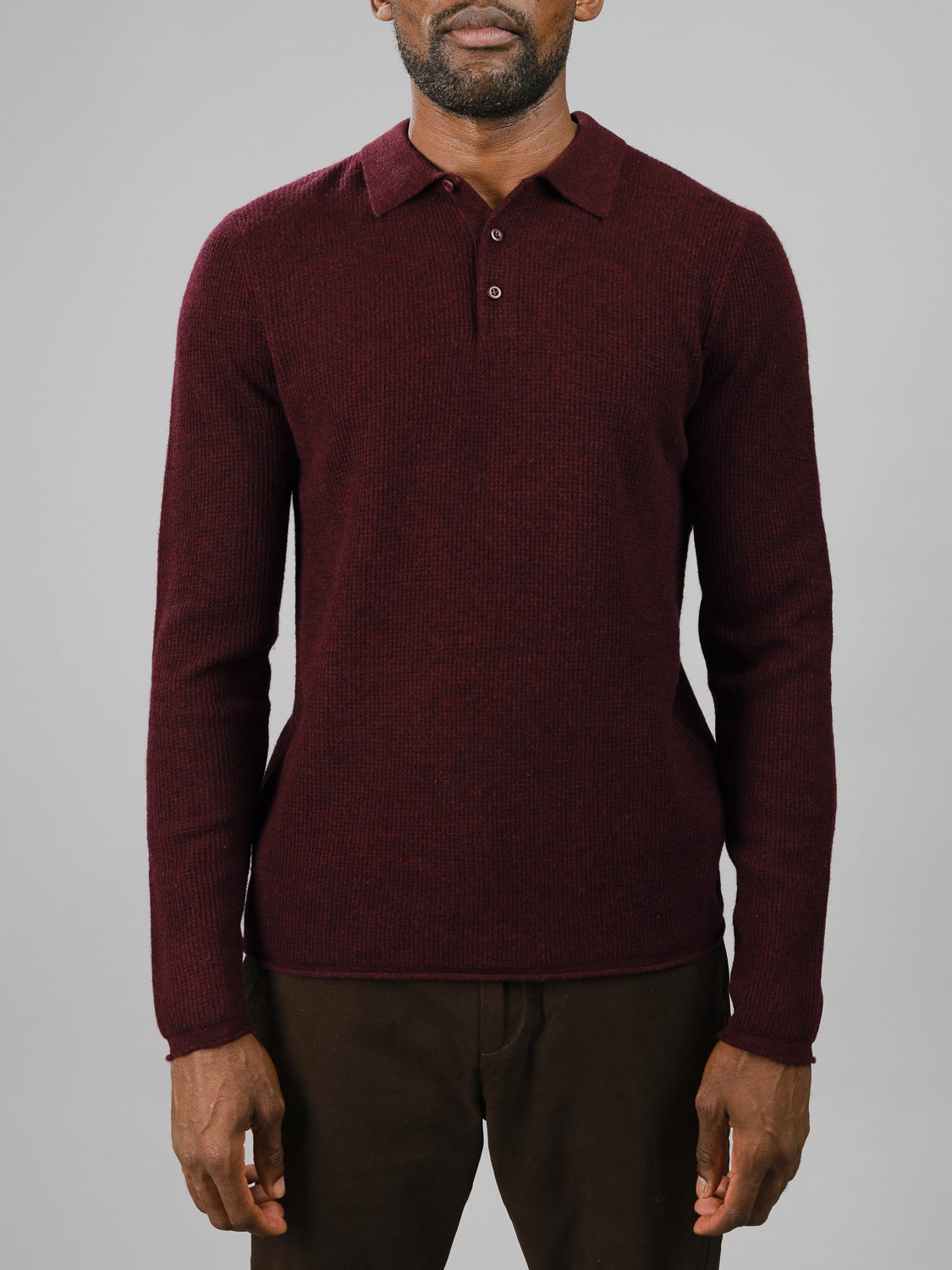 Wearing the Gothenberg Merino Sweater Polo by ID Menswear, a person exudes casual elegance in a dark red, collared, and buttoned long-sleeve knit sweater made of merino wool. The snug fit contrasts against a plain light gray background as they avoid facing the camera directly.