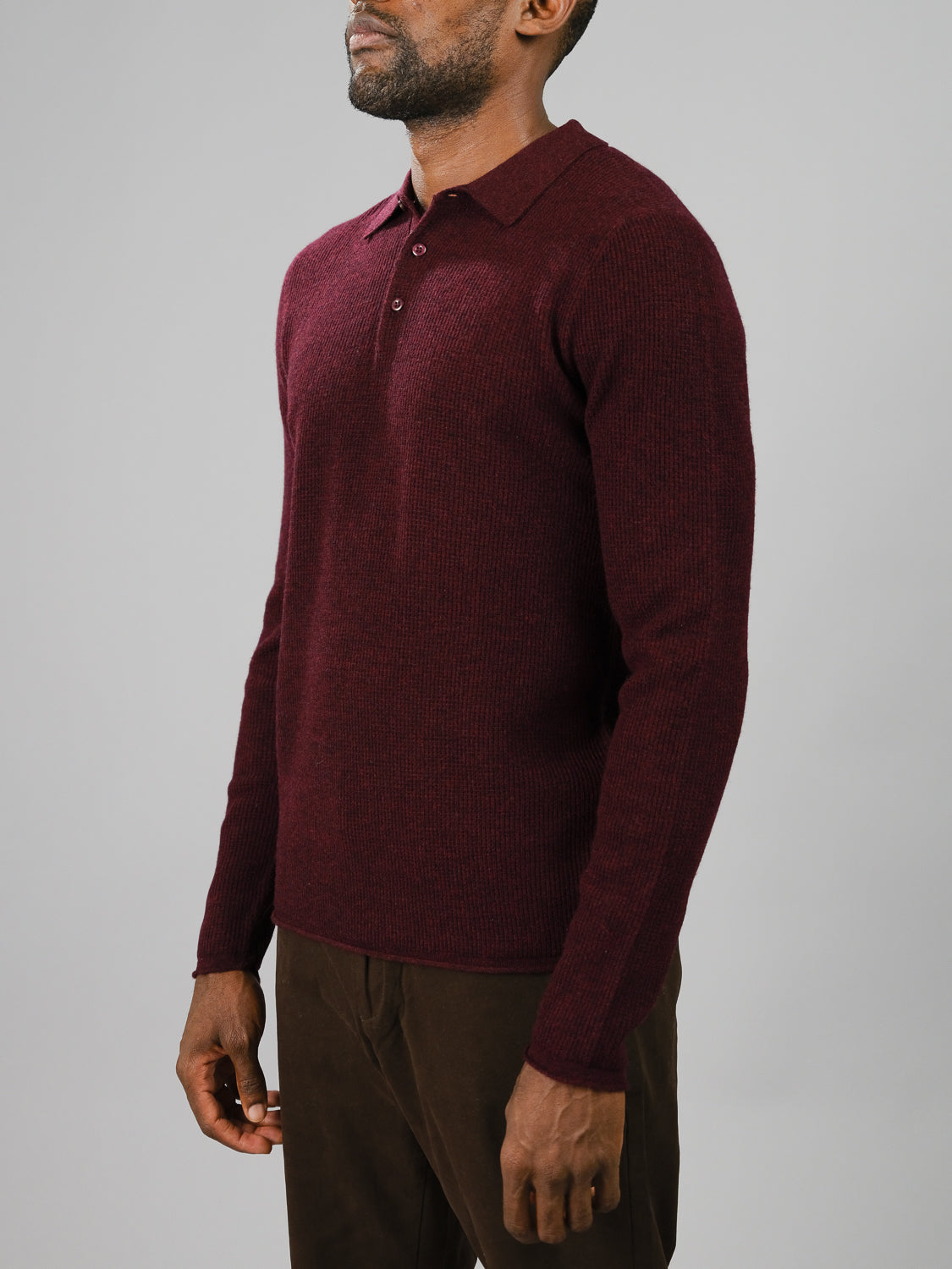 Wearing the Gothenberg Merino Sweater Polo from ID Menswear in dark red, a person embodies casual elegance with long sleeves. Paired with brown pants against a plain backdrop, the image is cropped from the shoulders down to maintain facial privacy.