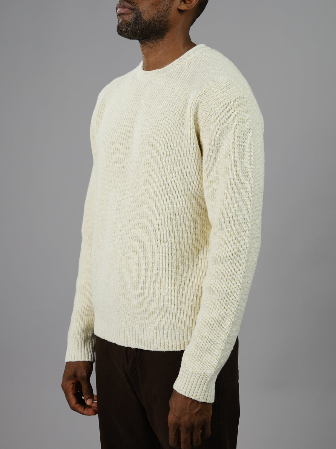 A man in a cream-colored Dublin Slub Cotton Rolled Neck Sweater from ID Menswear stands against a gray backdrop, his arms at his sides, embracing the cooler weather. Only part of his face is visible.