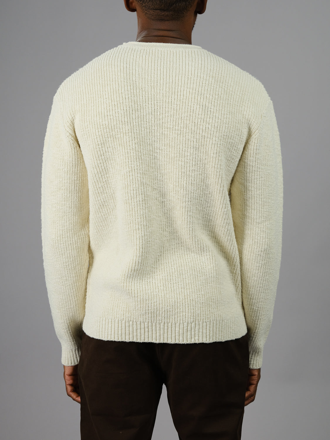 A person stands with their back to the camera, wrapped in an ID Menswear Dublin Slub Cotton Rolled Neck Sweater in cream ribbed knit, ideal for cooler days, stylishly paired with dark trousers against a plain gray backdrop.