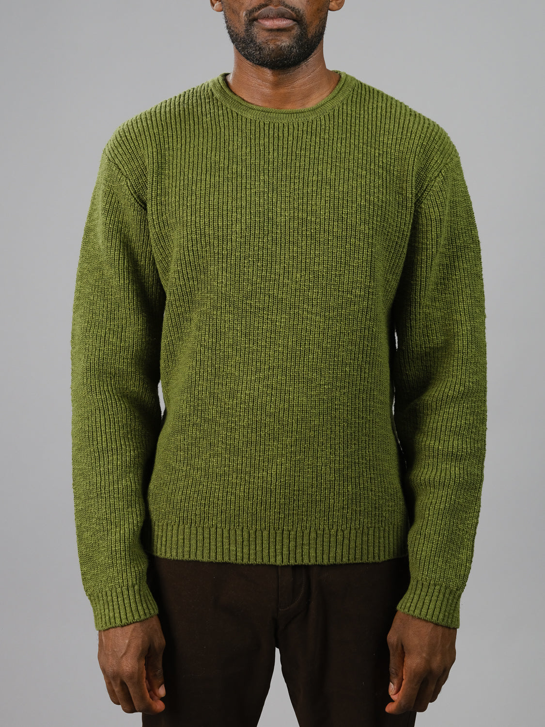 A person in an ID Menswear Dublin Slub Cotton Rolled Neck Sweater, ideal for cooler weather with its ribbed texture and long sleeves, stands against a plain gray background. Their head is partially out of frame, and theyre paired with dark pants. The sweater is thick and green.