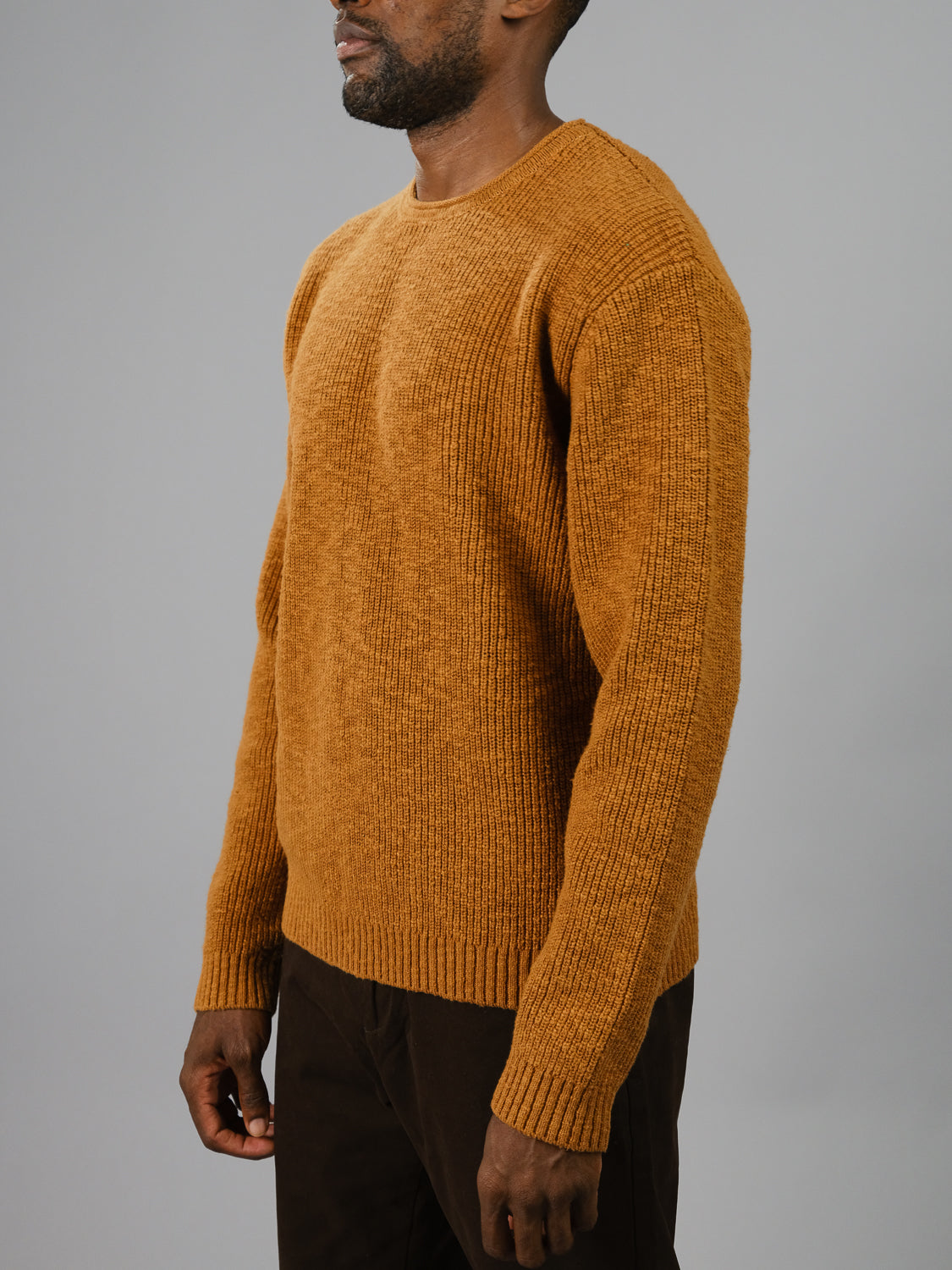 A person in a mustard Dublin Slub Cotton Rolled Neck Sweater from ID Menswear, featuring a ribbed texture and long sleeves, stands side view against a plain gray background with relaxed arms and dark pants, ideal for cooler weather.