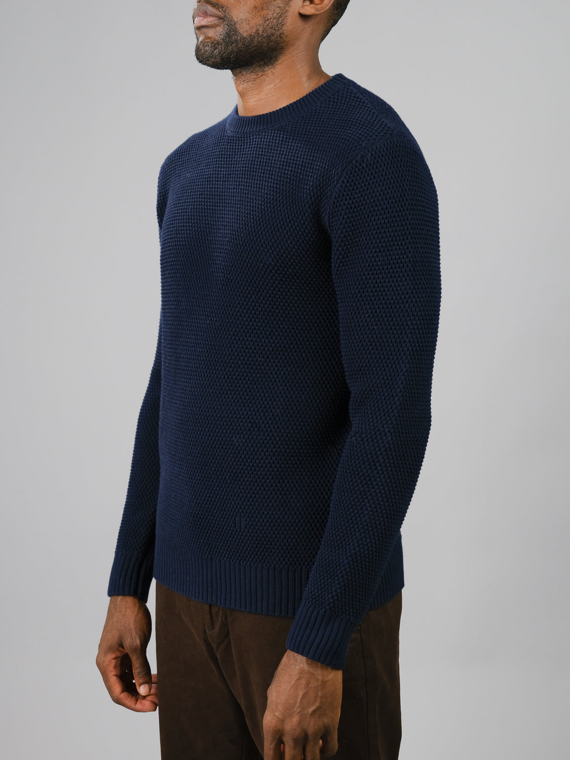 A man in a cozy and stylish Quebec Chunky Sweater by ID Menswear, featuring navy blue texture and Moss knit pattern, stands against a gray background. Facing slightly right, he pairs it with brown pants; the round neckline and ribbed cuffs enhance its appeal.