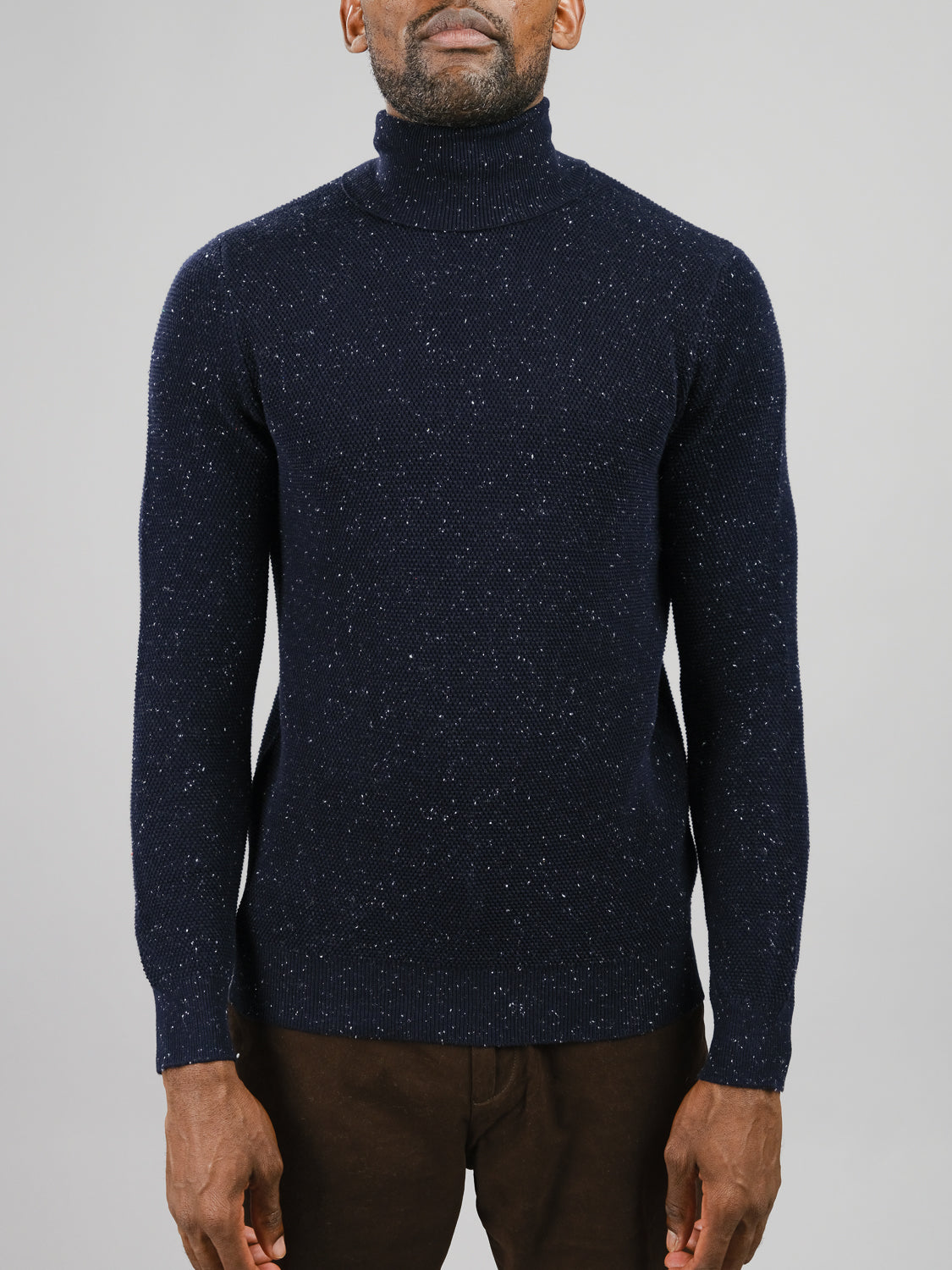 A person is wearing an ID Menswear Brampton High Neck Cotton Sweater in dark blue with speckled yarn and brown pants. The high neck design elegantly contrasts the plain gray background.