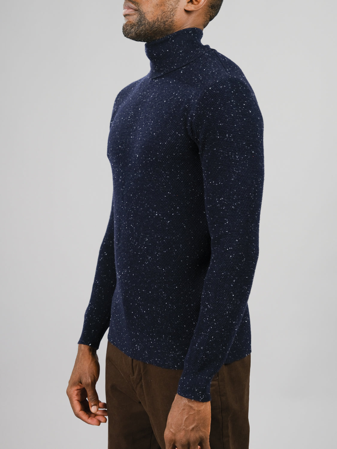 A man in an ID Menswear Brampton High Neck Cotton Sweater with a speckled yarn design and brown pants stands sideways against a plain gray backdrop.
