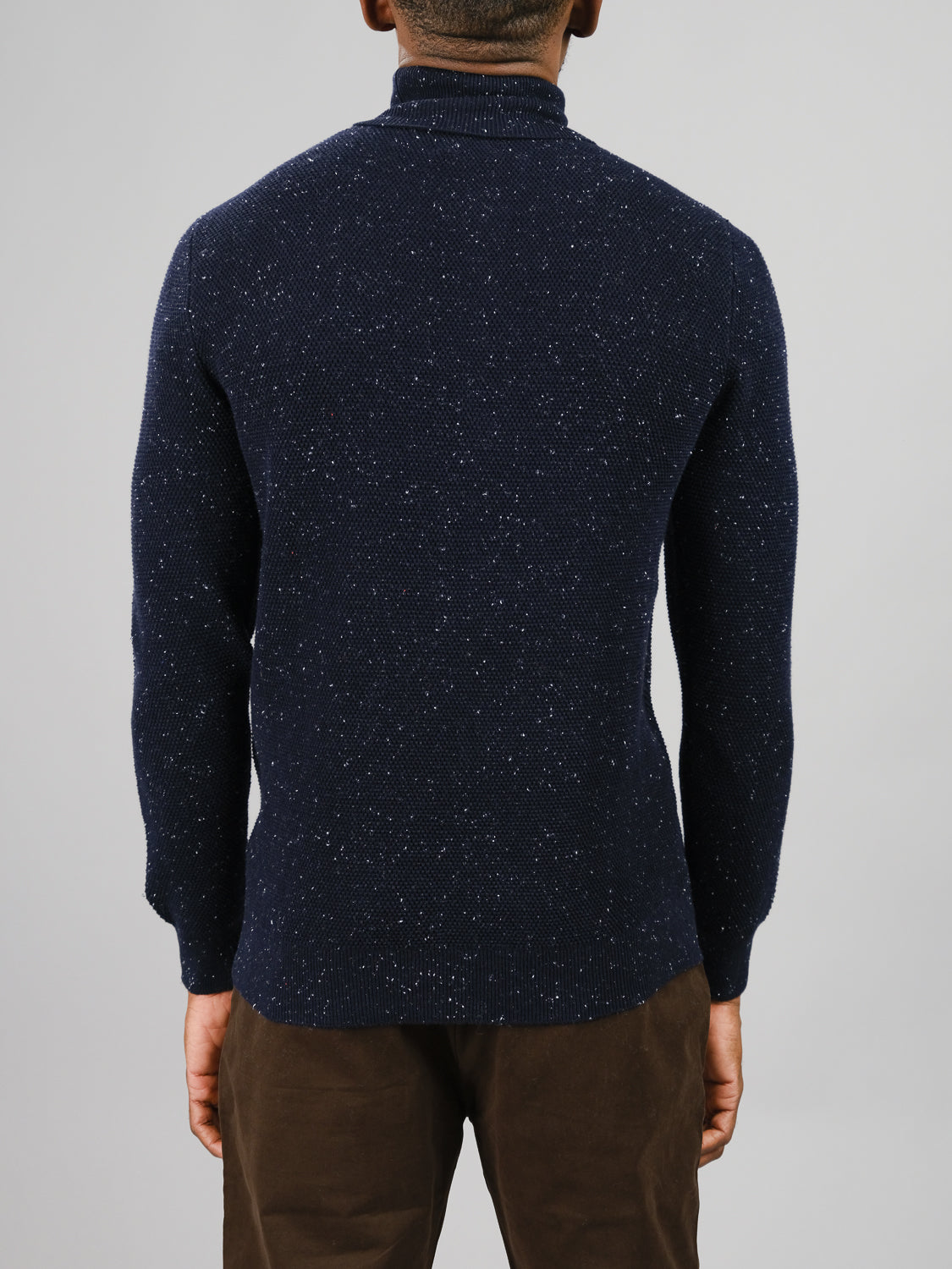A person is seen from behind wearing a dark blue Brampton High Neck Cotton Sweater by ID Menswear, made of speckled yarn, and dark brown pants against a light gray background.