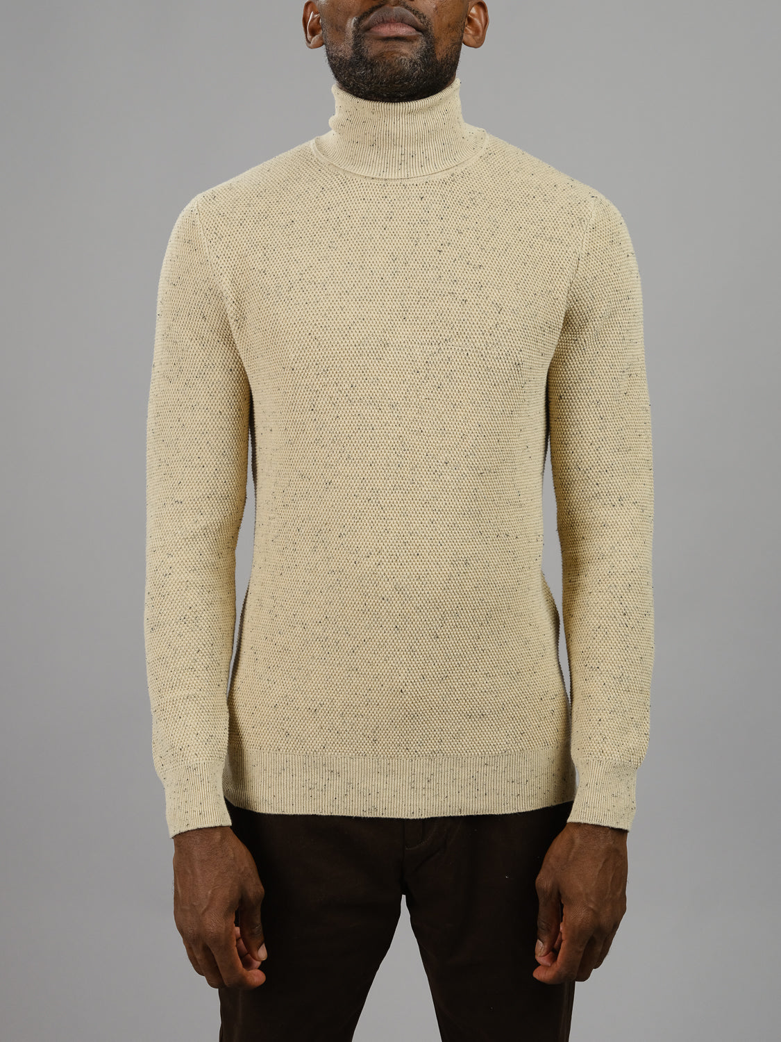 A person wearing an ID Menswear Brampton High Neck Cotton Sweater, featuring a light beige design and textured knit pattern, stands against a plain gray background with dark pants. The face is not visible.