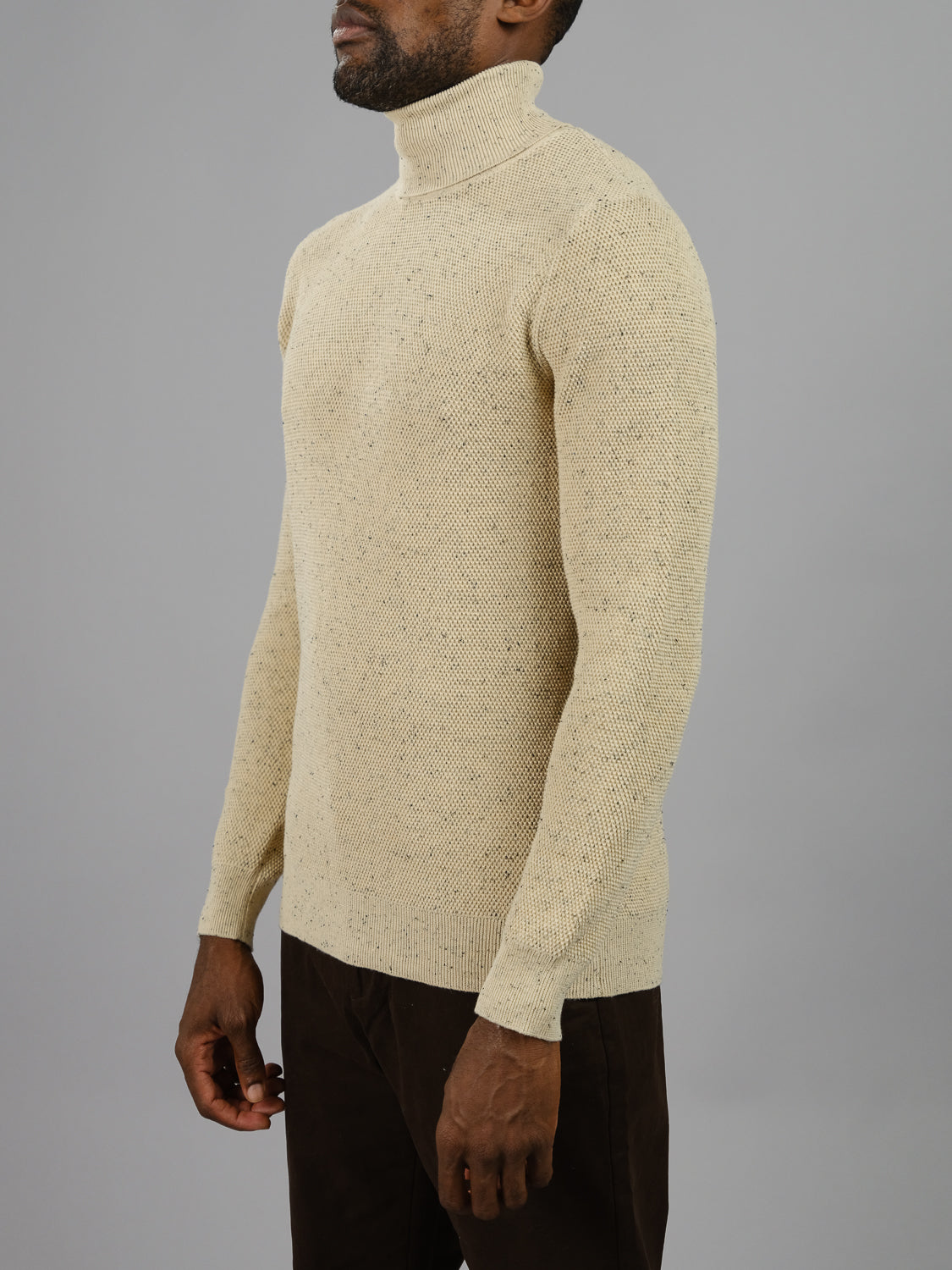 A man wears a cream ID Menswear Brampton High Neck Cotton Sweater with dark pants against a gray background, captured from the side with relaxed arms.
