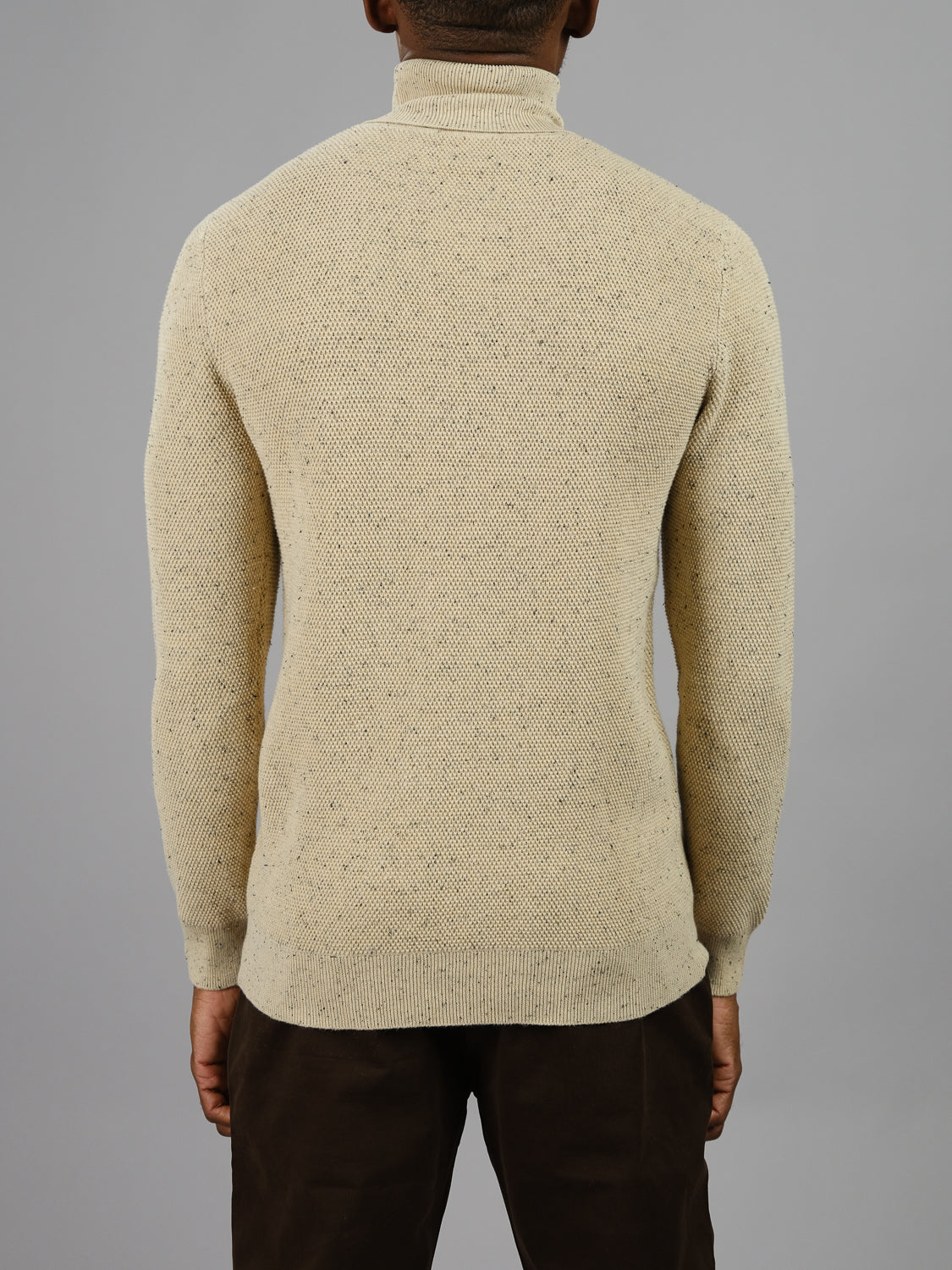 A person stands against a plain gray background, wearing ID Menswears Brampton High Neck Cotton Sweater in cream with speckled yarn texture, paired with brown pants.