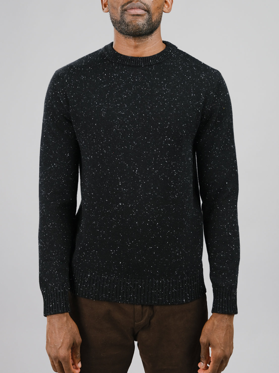 A person in a dark Malmo Speckled Cotton Sweater by ID Menswear is against a plain gray backdrop. Their hands are relaxed at their sides, and they wear brown pants. This crew neck sweater has ribbed collar, cuffs, and hem.