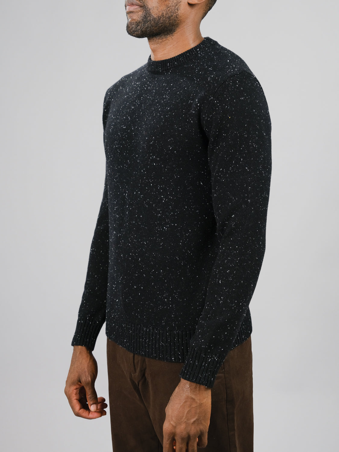 A person wearing an ID Menswear Malmo Speckled Cotton Sweater in black speckled design paired with brown pants stands against a plain gray background. The crew neck sweater has ribbed cuffs and hem, and they are shown from the chest down, facing slightly left.