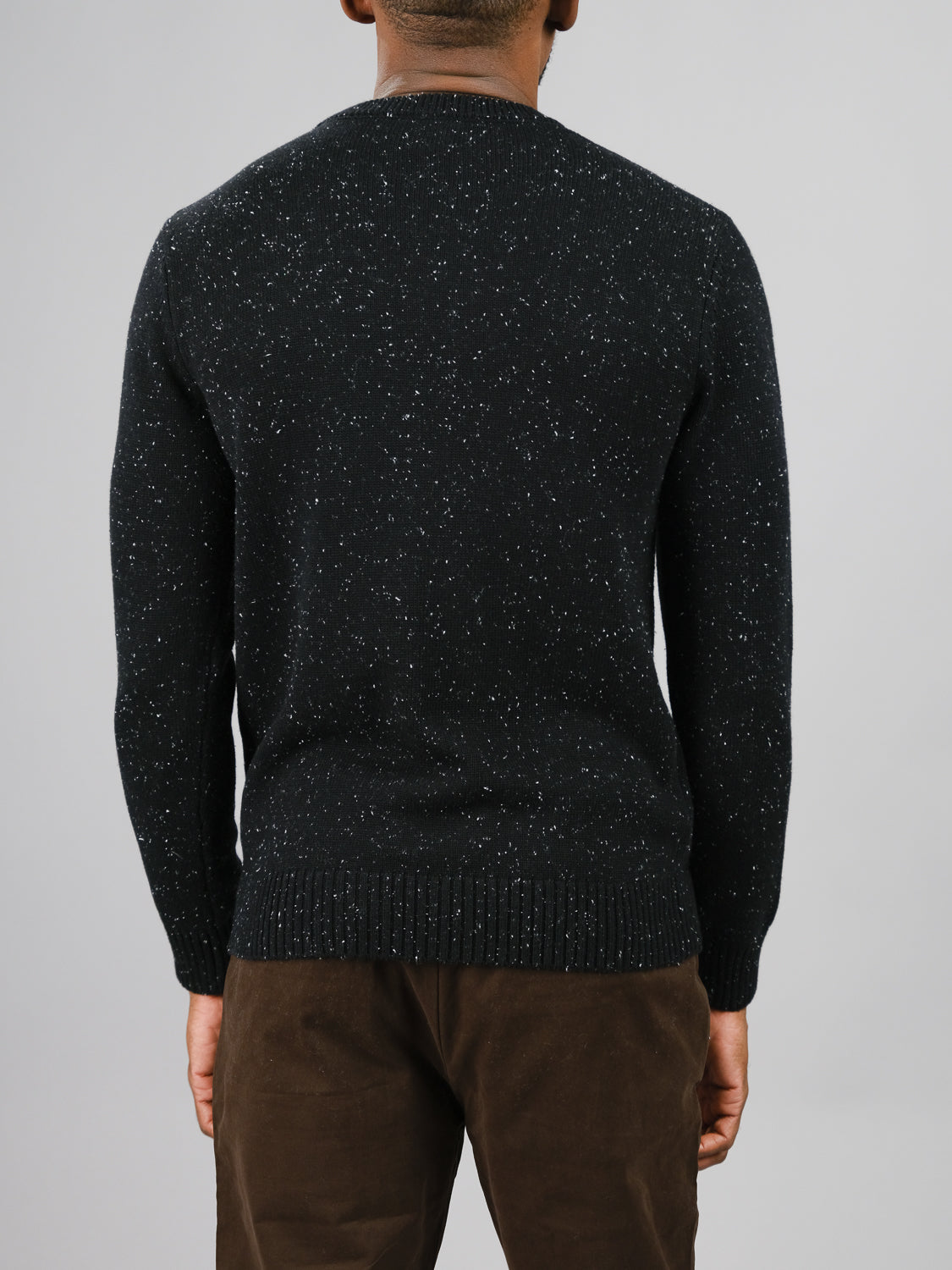 A person is seen from behind in a dark speckled Malmo Speckled Cotton Sweater by ID Menswear and brown pants, set against a light gray backdrop.