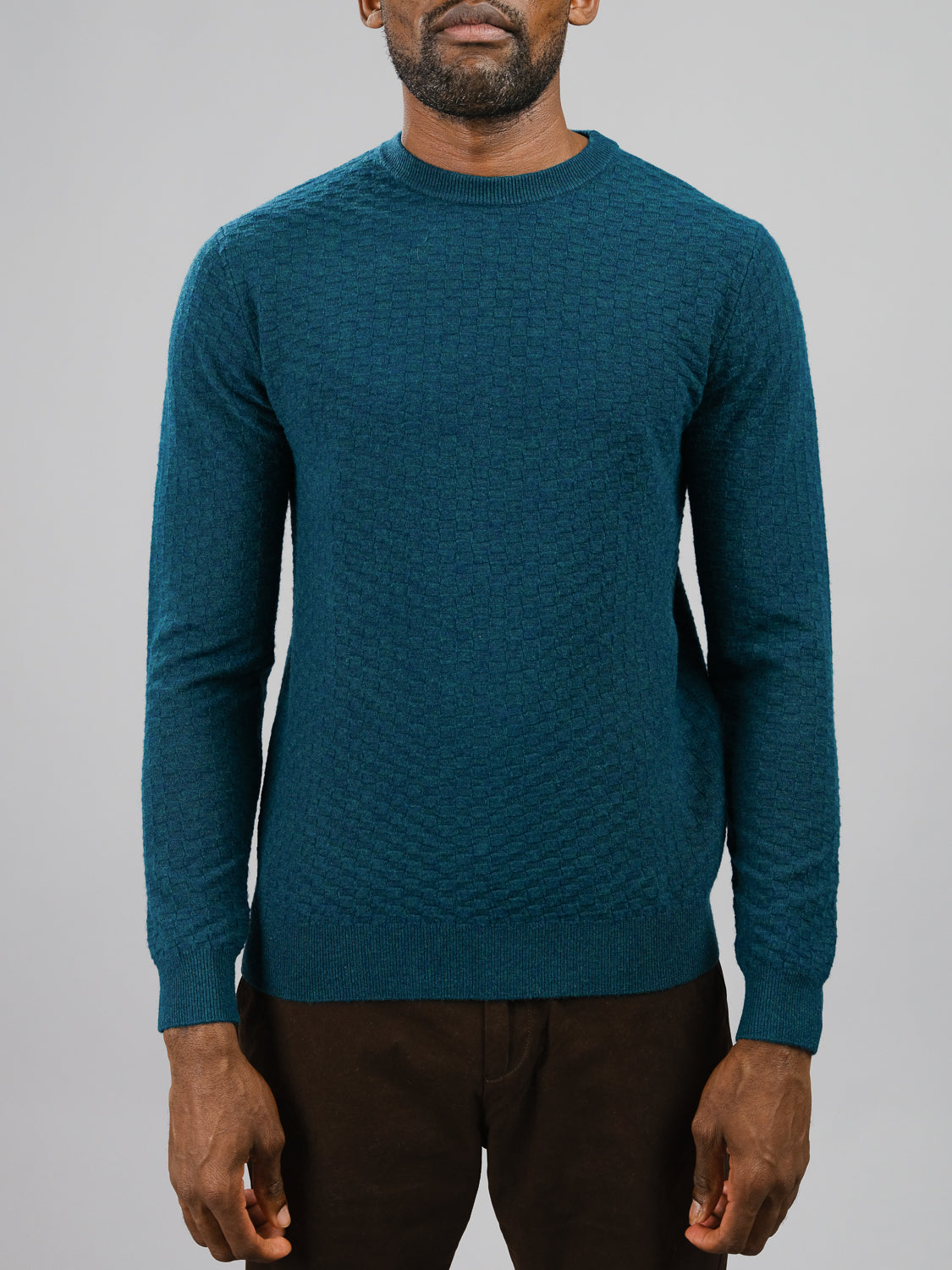 A person in ID Menswears Stockholm Merino Wool Sweater, a classic crewneck in teal with a subtle checkered pattern, and dark brown pants stands against a plain gray background. The lightweight sweater hangs relaxed as the persons arms rest by their sides.