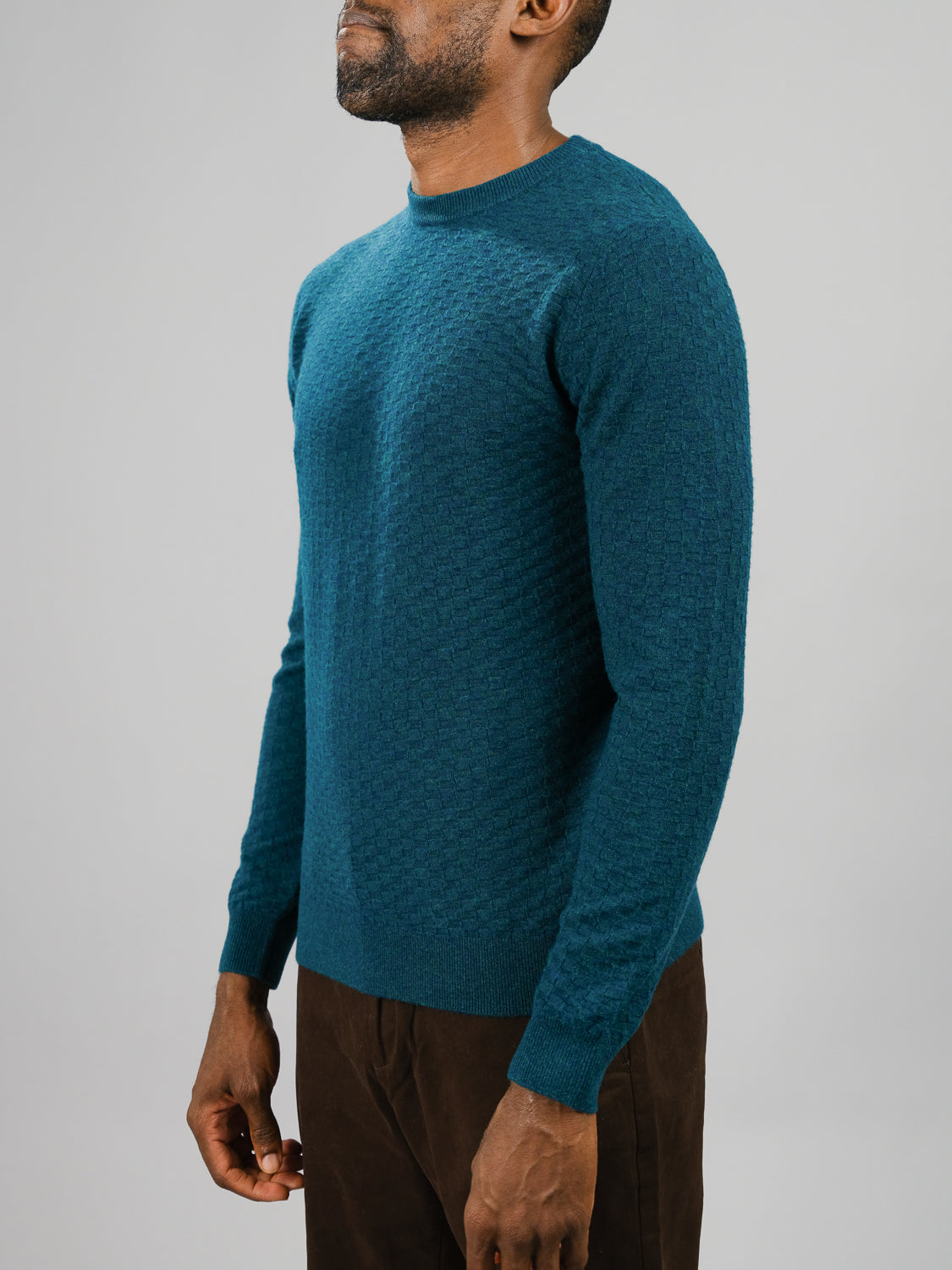 A man in a Stockholm Merino Wool Sweater from ID Menswear, featuring a textured teal design, pairs it with brown pants as he stands against a plain gray background, facing slightly to the side.