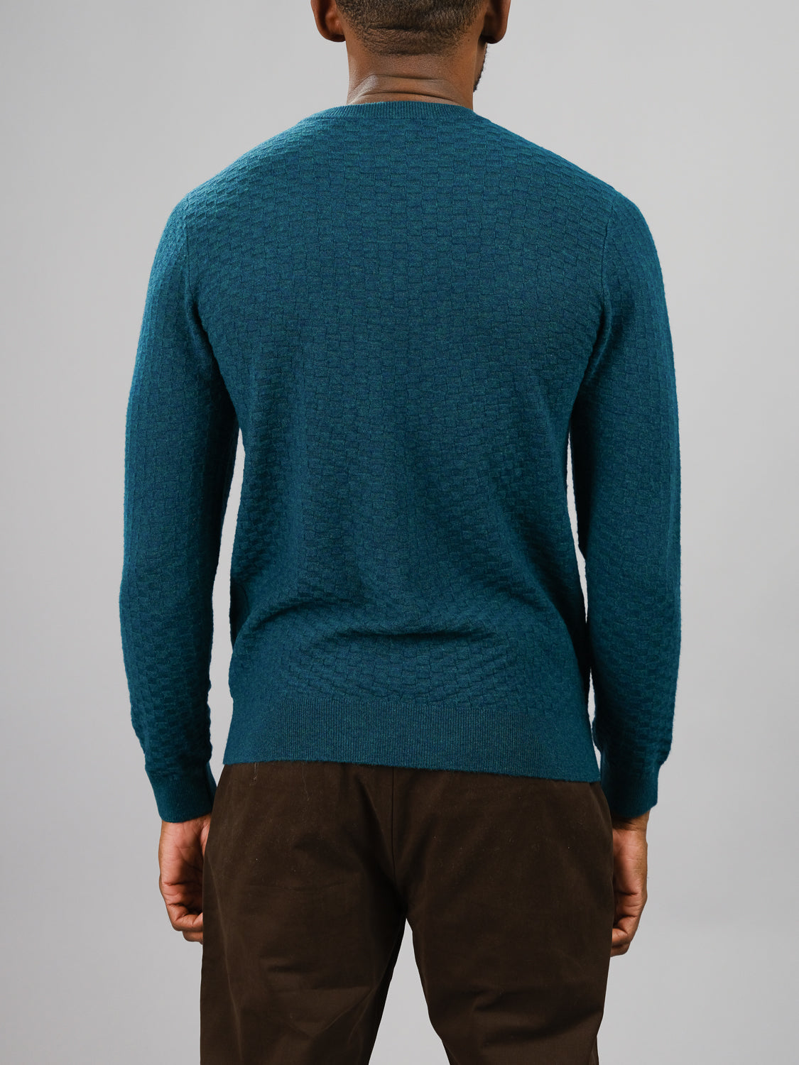 A person in an ID Menswear Stockholm Merino Wool Sweater, featuring a textured blue-green design, and brown pants is shown from the back against a plain gray background.