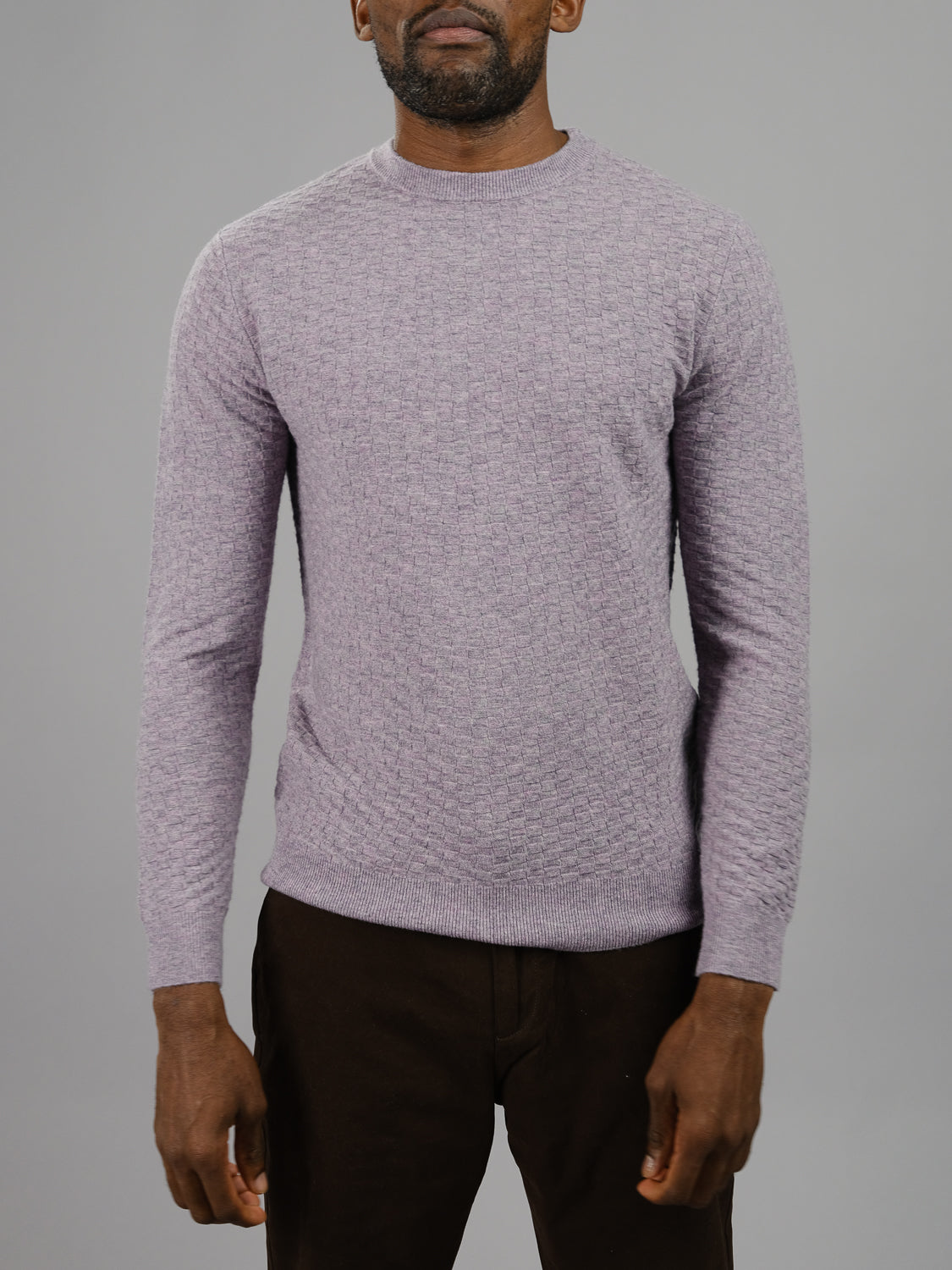 A person in a Stockholm Merino Wool Sweater by ID Menswear, featuring a lightweight texture, light purple hue, classic crewneck, and subtle pattern with long sleeves, stands against a plain gray background in dark pants with relaxed hands by their sides.