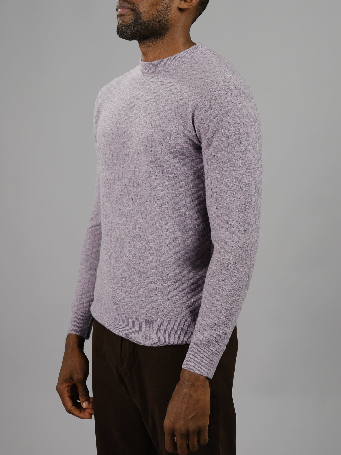 A person donning a Stockholm Merino Wool Sweater in light purple from ID Menswear and dark brown pants stands against a gray background. The lightweight sweater drapes elegantly as they slightly face left, hands relaxed by their sides.