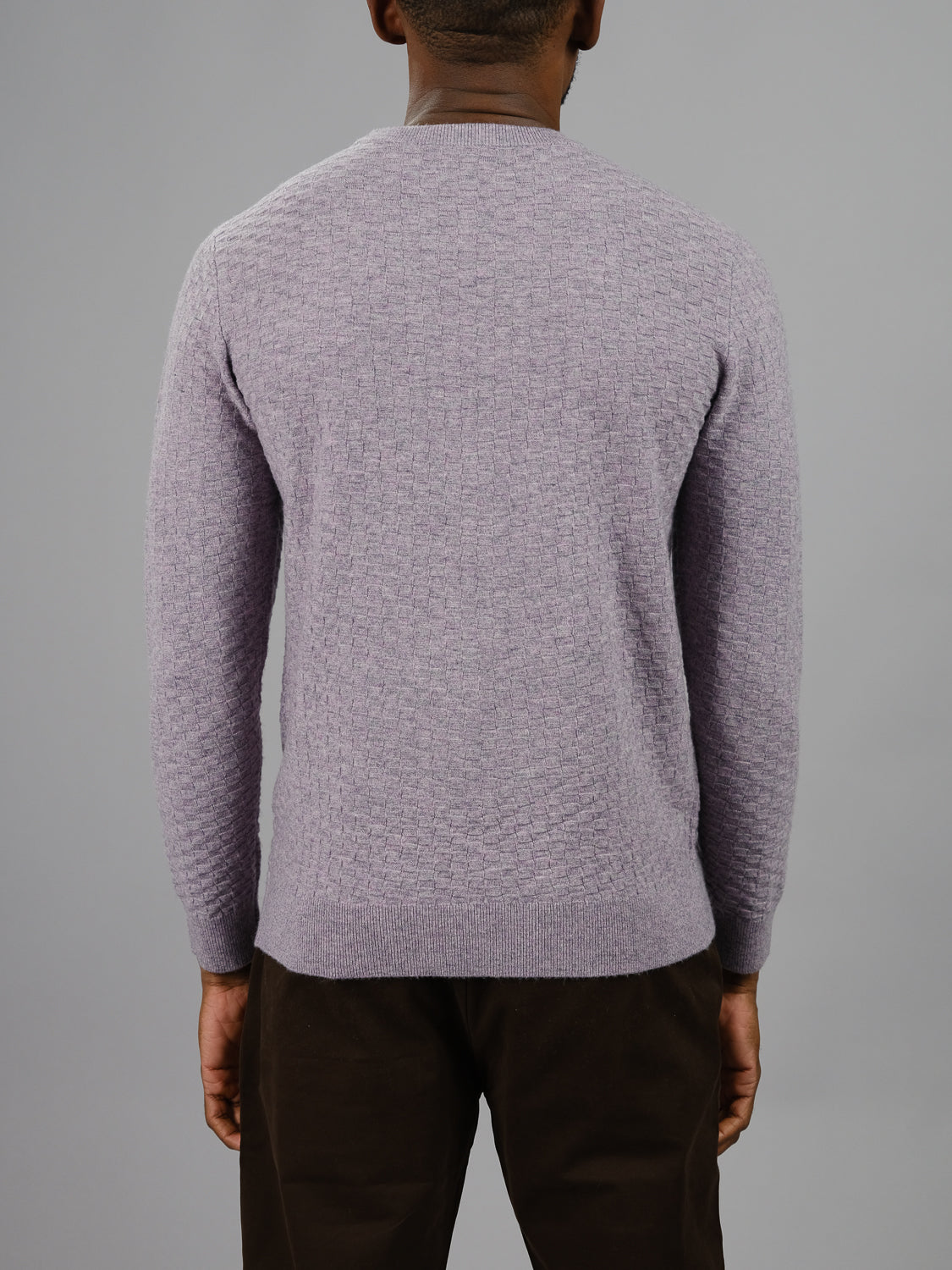 A person in an ID Menswear Stockholm Merino Wool Sweater, featuring a light purple hue and classic crewneck, pairs it with dark pants. They pose against a gray backdrop, highlighting the sweaters lightweight texture from behind.