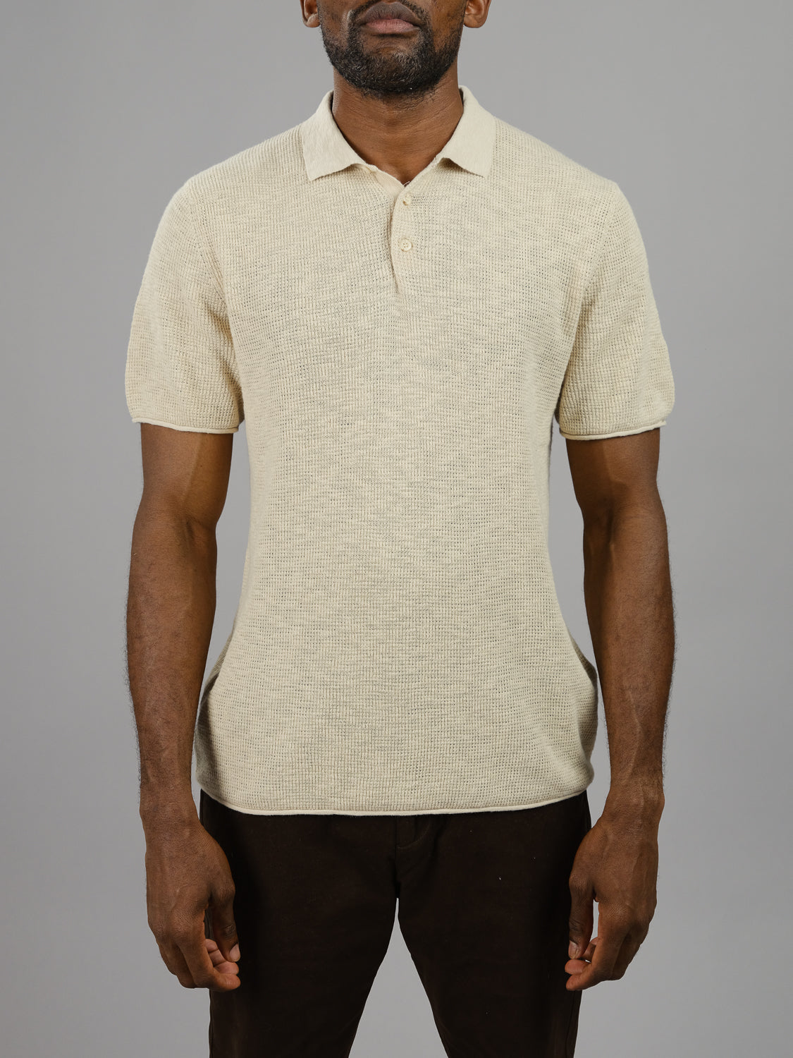 A man wearing the Athens Waffle Knit Polo from ID Menswear, paired with dark pants, stands against a plain gray background, exuding a relaxed and casual style with his hands at his sides.