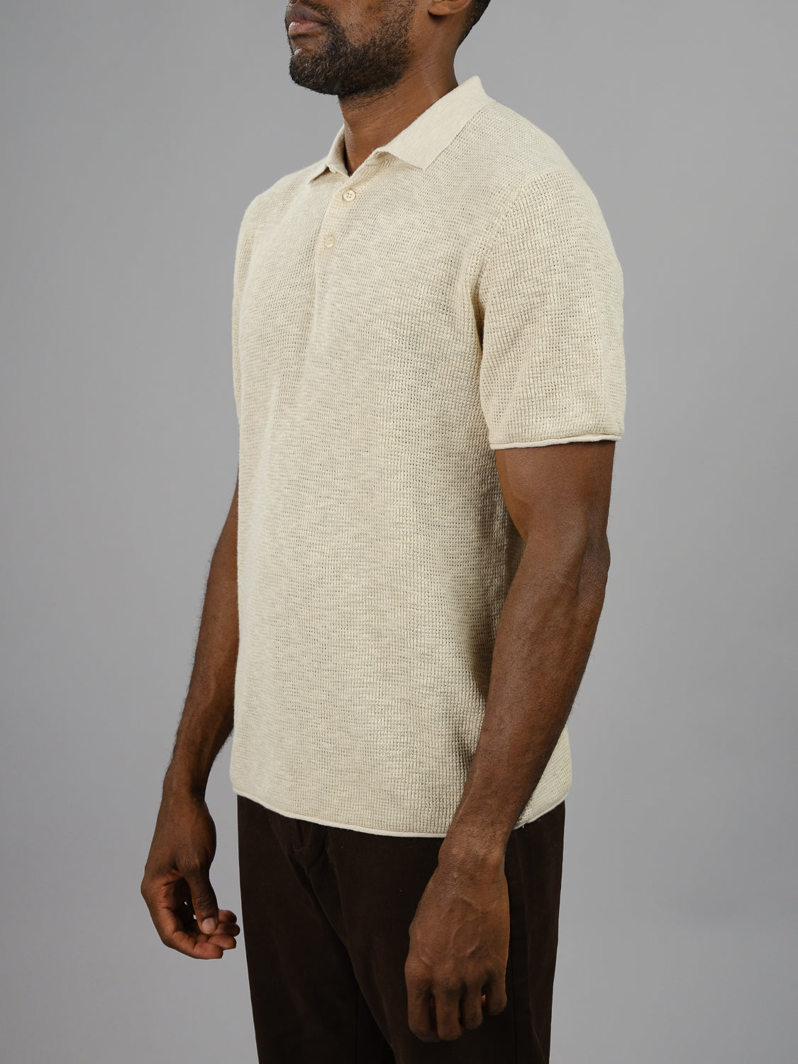 A man in the Athens Waffle Knit Polo by ID Menswear, beige with short sleeves, pairs it with dark brown pants. He stands against a plain gray background, his arms relaxed by his sides as he turns slightly left, showcasing his casual style and the textured look of his outfit.