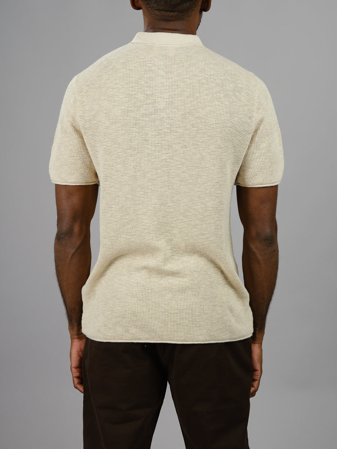 A person is seen from the back wearing ID Menswears Athens Waffle Knit Polo in light beige with dark pants against a gray background, arms at their sides, highlighting the shirt’s subtle texture and casual style.