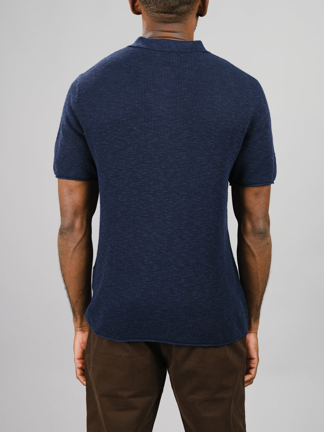 Rear view of a person in the Athens Waffle Knit Polo by ID Menswear, a navy blue, short-sleeved shirt, paired with brown pants for a textured look against a plain light gray background.