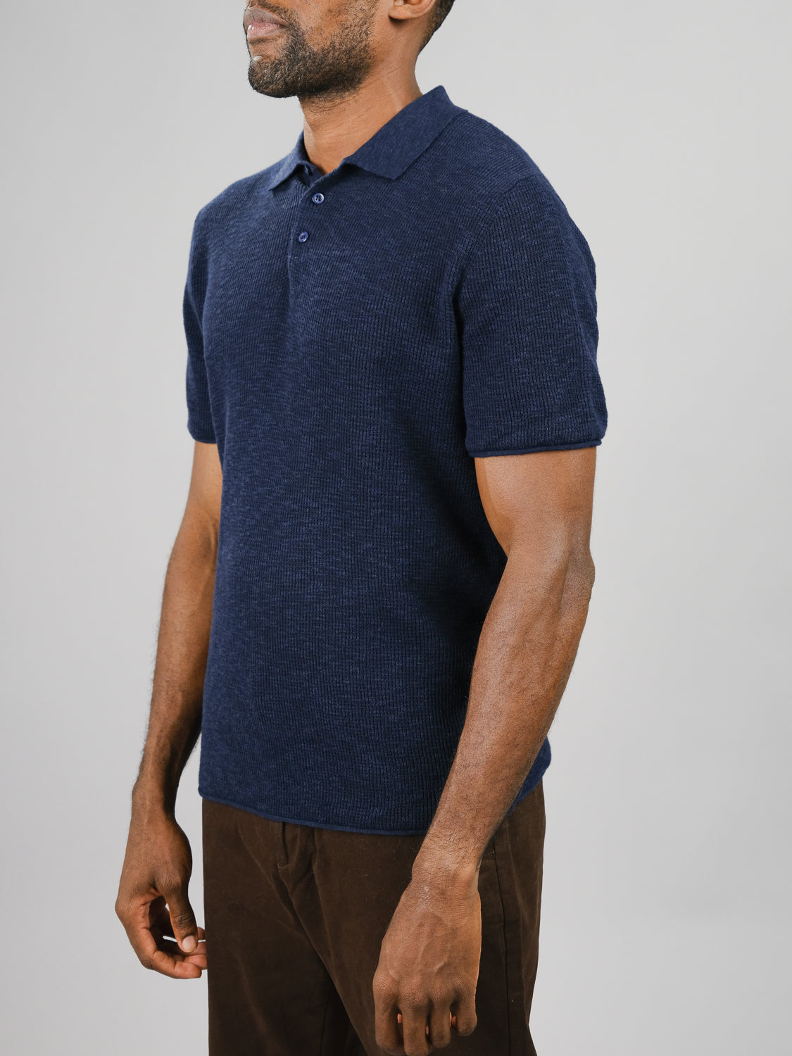 A person wearing the Athens Waffle Knit Polo from ID Menswear in navy blue and brown pants stands against a plain background. The textured polo, with its collar buttons, projects a casual style. The persons face is not visible.