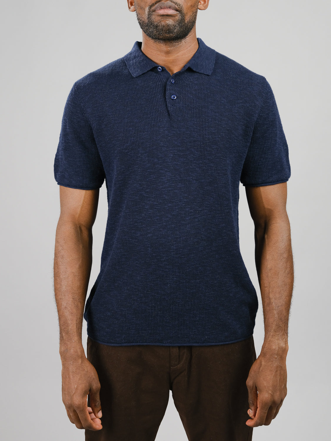 A man wears the Athens Waffle Knit Polo by ID Menswear in navy blue with brown pants against a light gray backdrop, emphasizing the textured design and color of the polo, capturing his casual style.