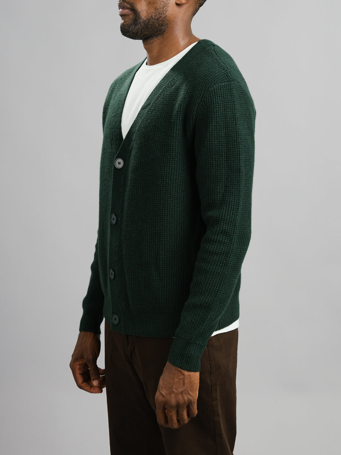 A man stands against a gray background wearing the ID Menswear Dundee Waffle Knit Merino Cardigan in dark green over a white t-shirt and brown pants. His face is partially out of frame, and his hands rest at his sides.