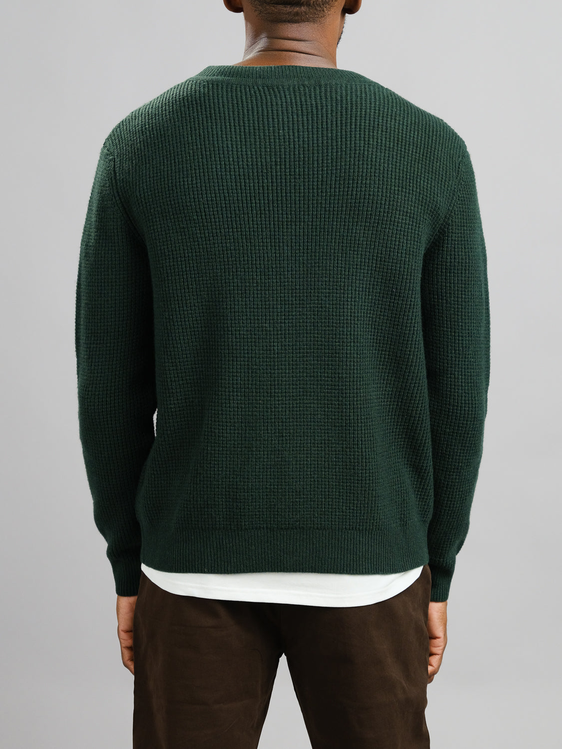 A person stands with their back to the camera, wearing the Dundee Waffle Knit Merino Cardigan by ID Menswear in dark green over a white shirt and dark pants, set against a plain light gray background.