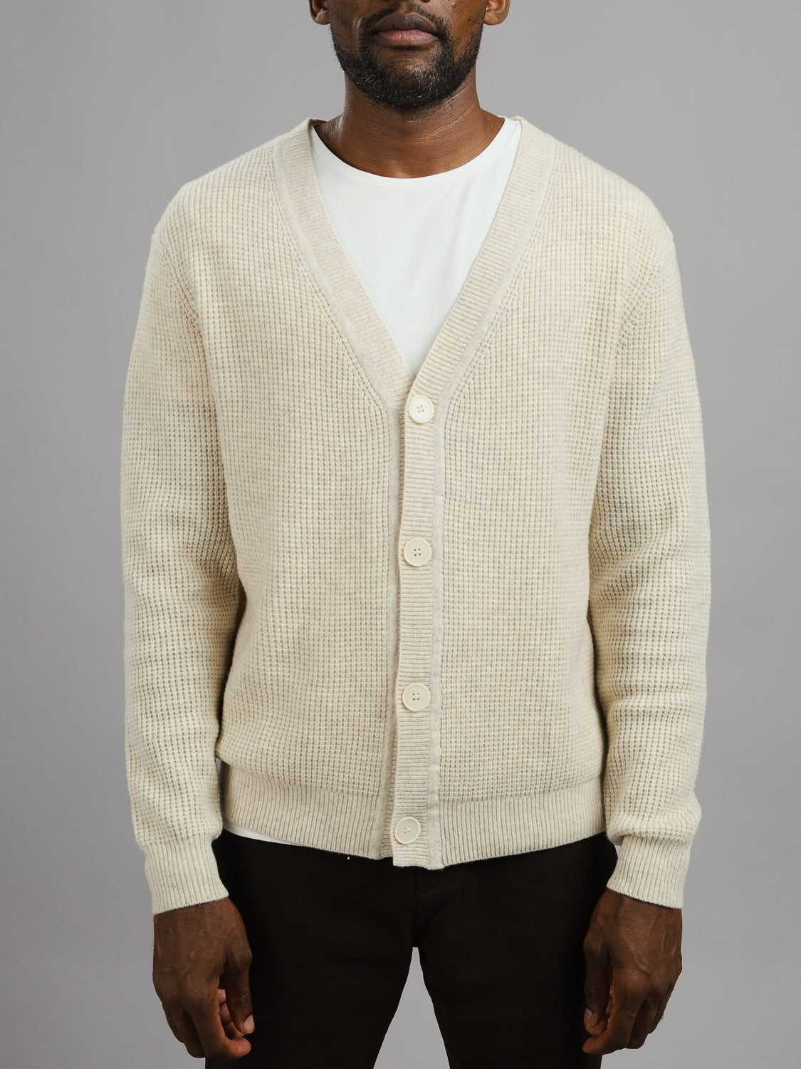 A man models the Dundee Waffle Knit Merino Cardigan from ID Menswear—cream-colored and buttoned over a white T-shirt with dark pants—standing against a plain gray background. The shot highlights the cardigan and upper body.