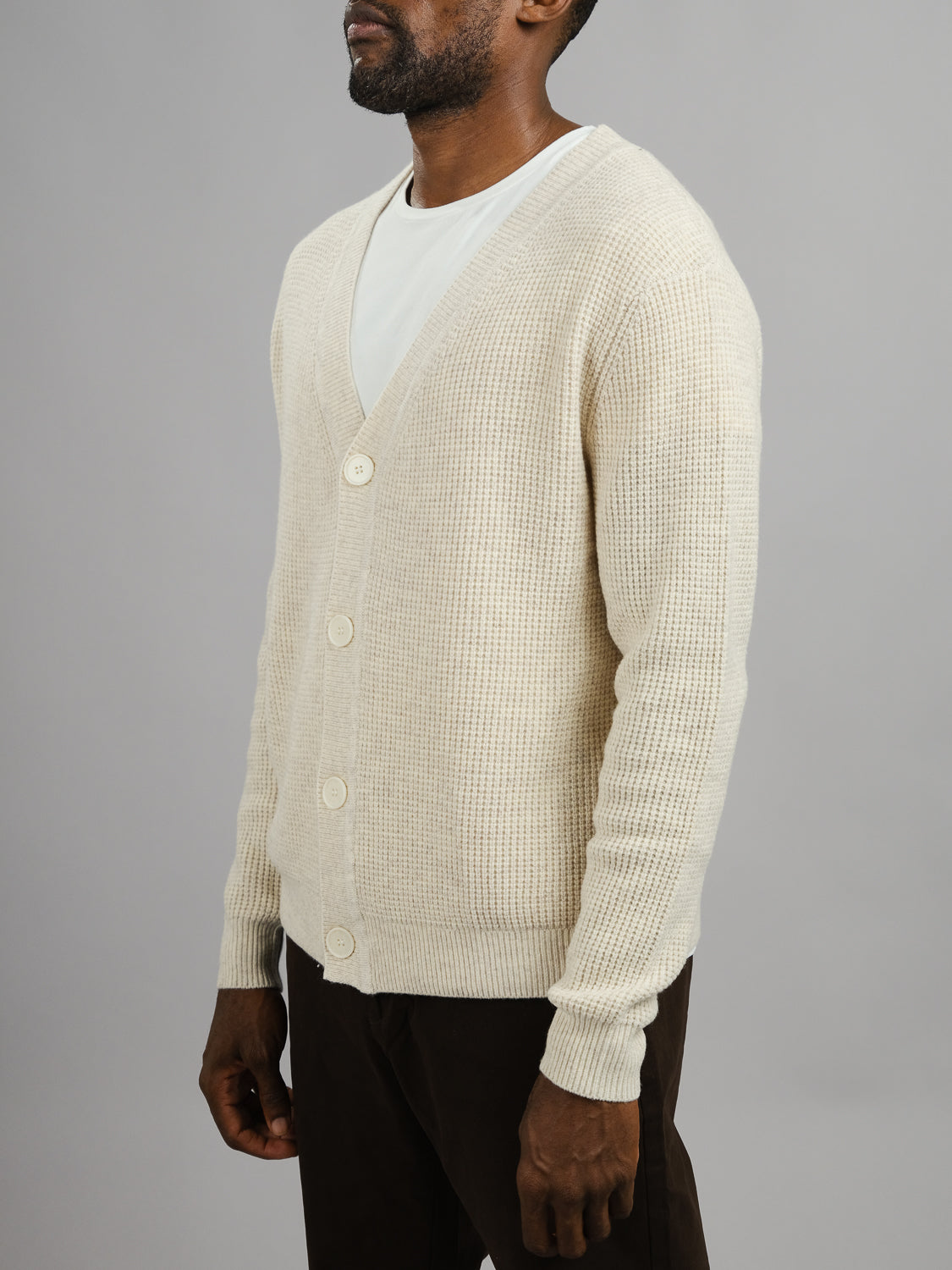 A man stands against a gray background, wearing the cream-colored Dundee Waffle Knit Merino Cardigan from ID Menswear over a white t-shirt and dark pants. His face is partially out of frame.