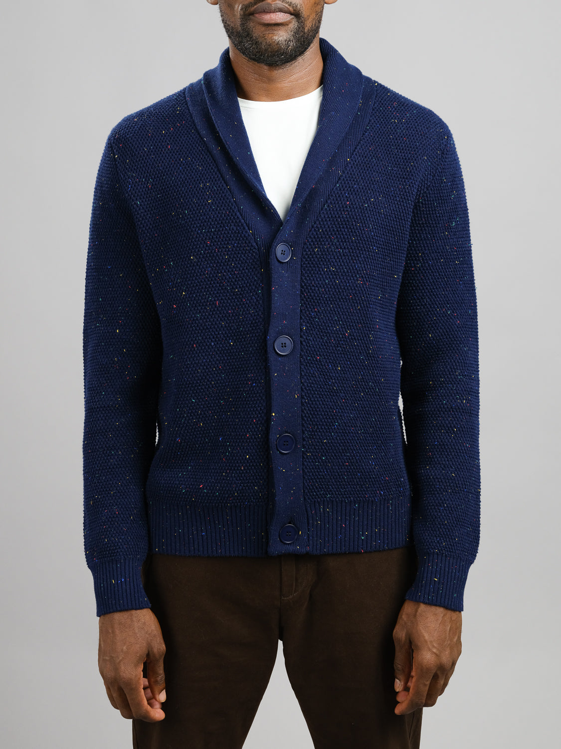 A person wears a dark blue Vermont Shawl Collar Cardigan by ID Menswear over a white shirt and brown pants. Crafted from speckled yarn, the cardigan features a textured knit pattern. The image focuses on the torso against a plain gray background.